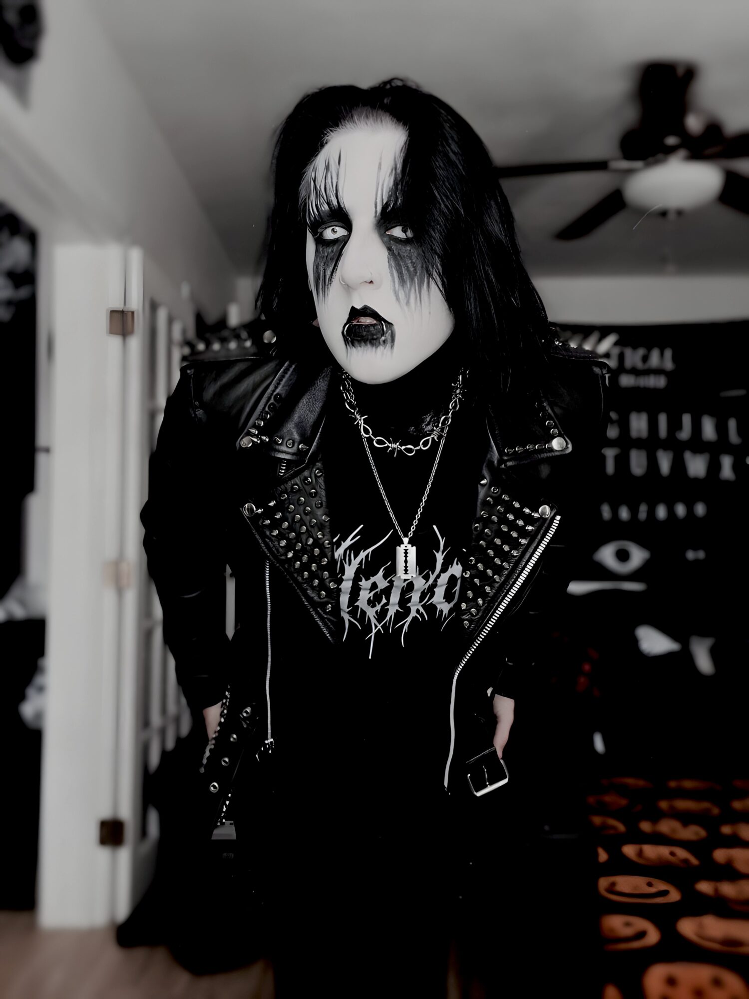Person with black and white makeup, black hair, wearing a leather jacket and chains, standing indoors.