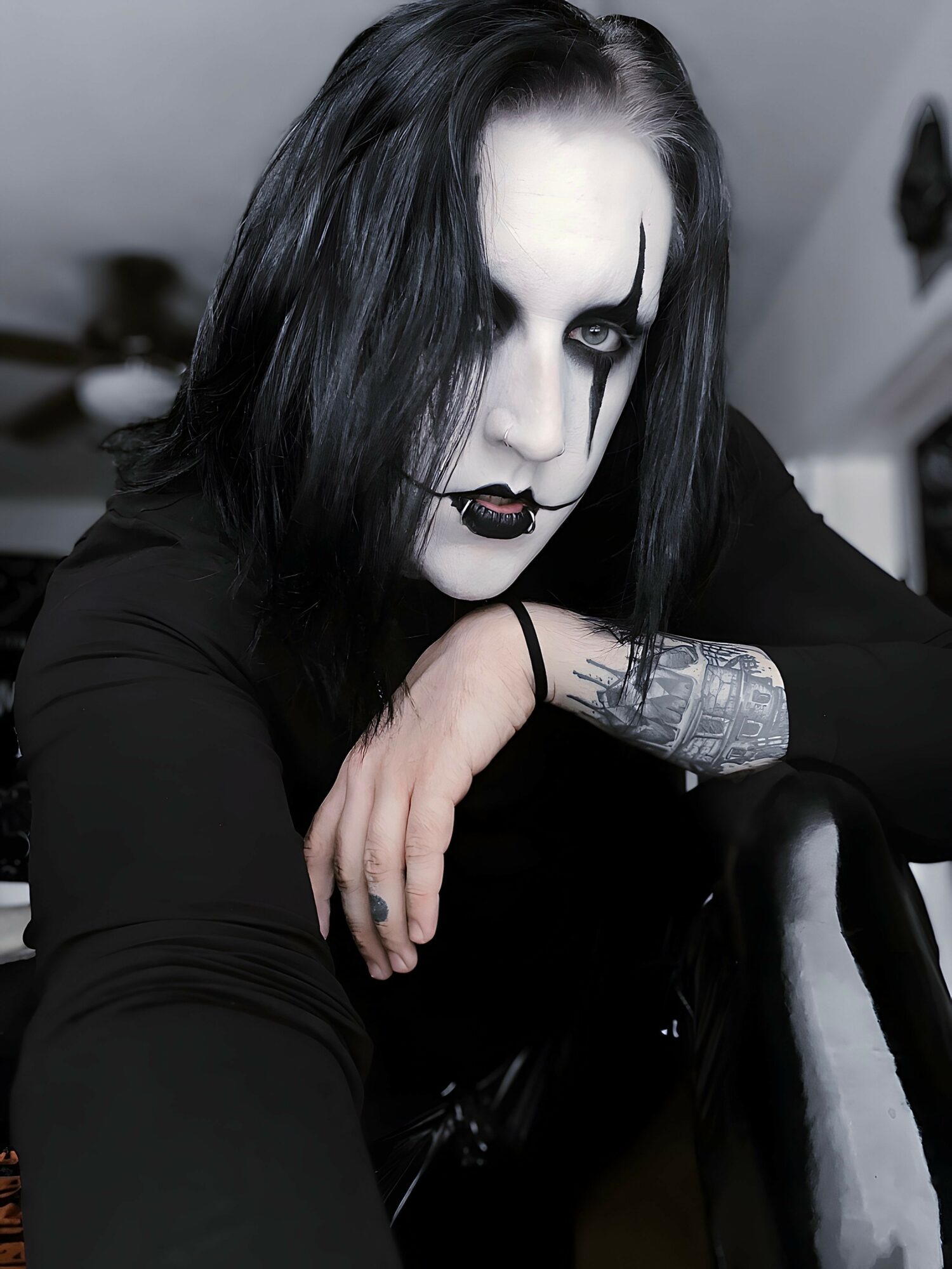 Person with black hair, white face makeup, dark eye makeup, and black lipstick, resting arm on knee, tattoo visible on wrist.