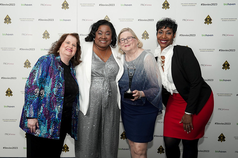 Four women standing together, smiling, in front of a backdrop with logos and text, dressed in formal attire.