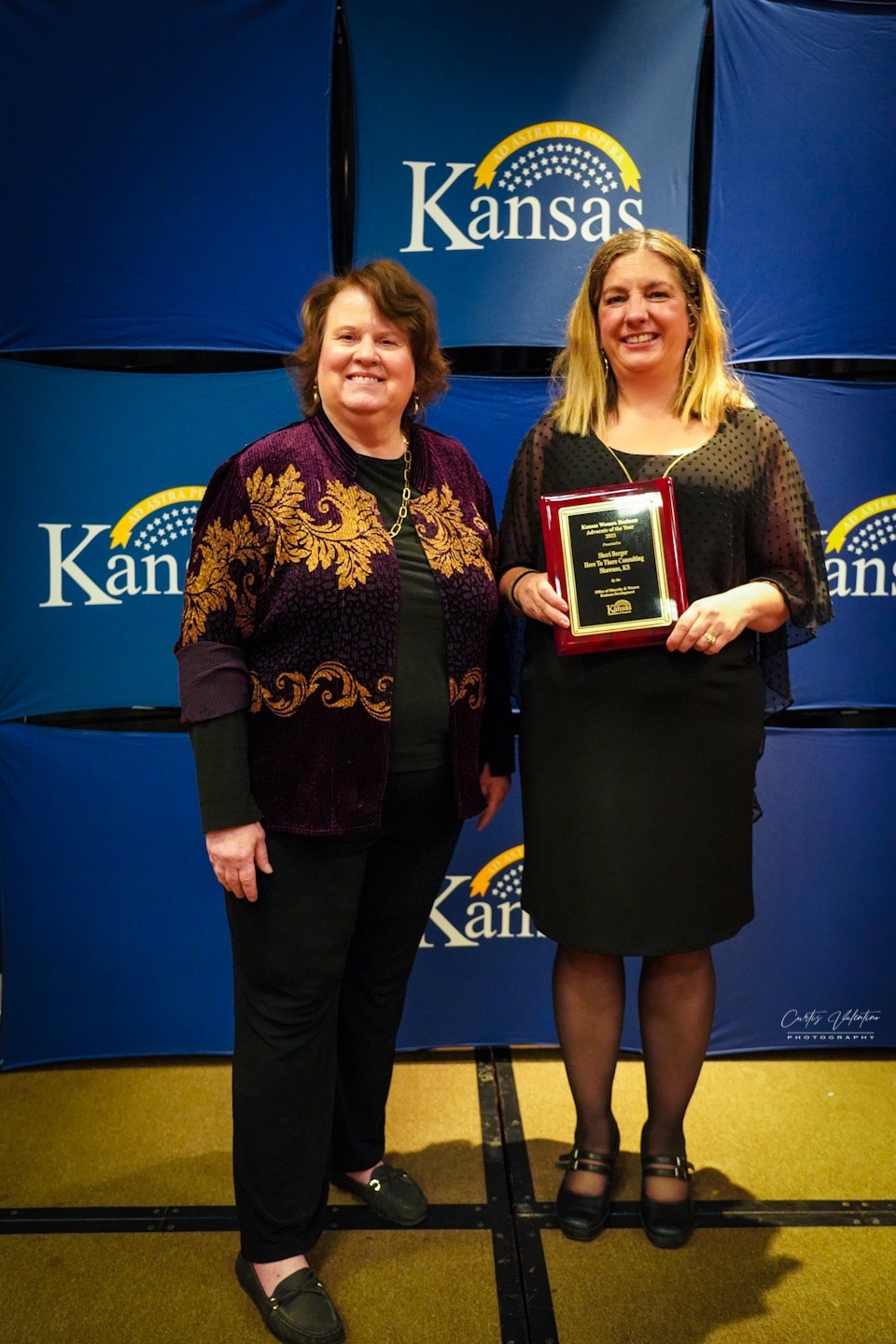 Two women standing in front of a Kansas backdrop, one holding an award plaque, smiling.