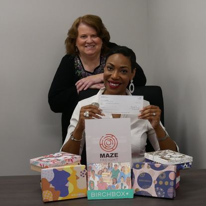 Two women smiling, one sitting and holding a check, the other standing behind, with boxes and a sign on the table.