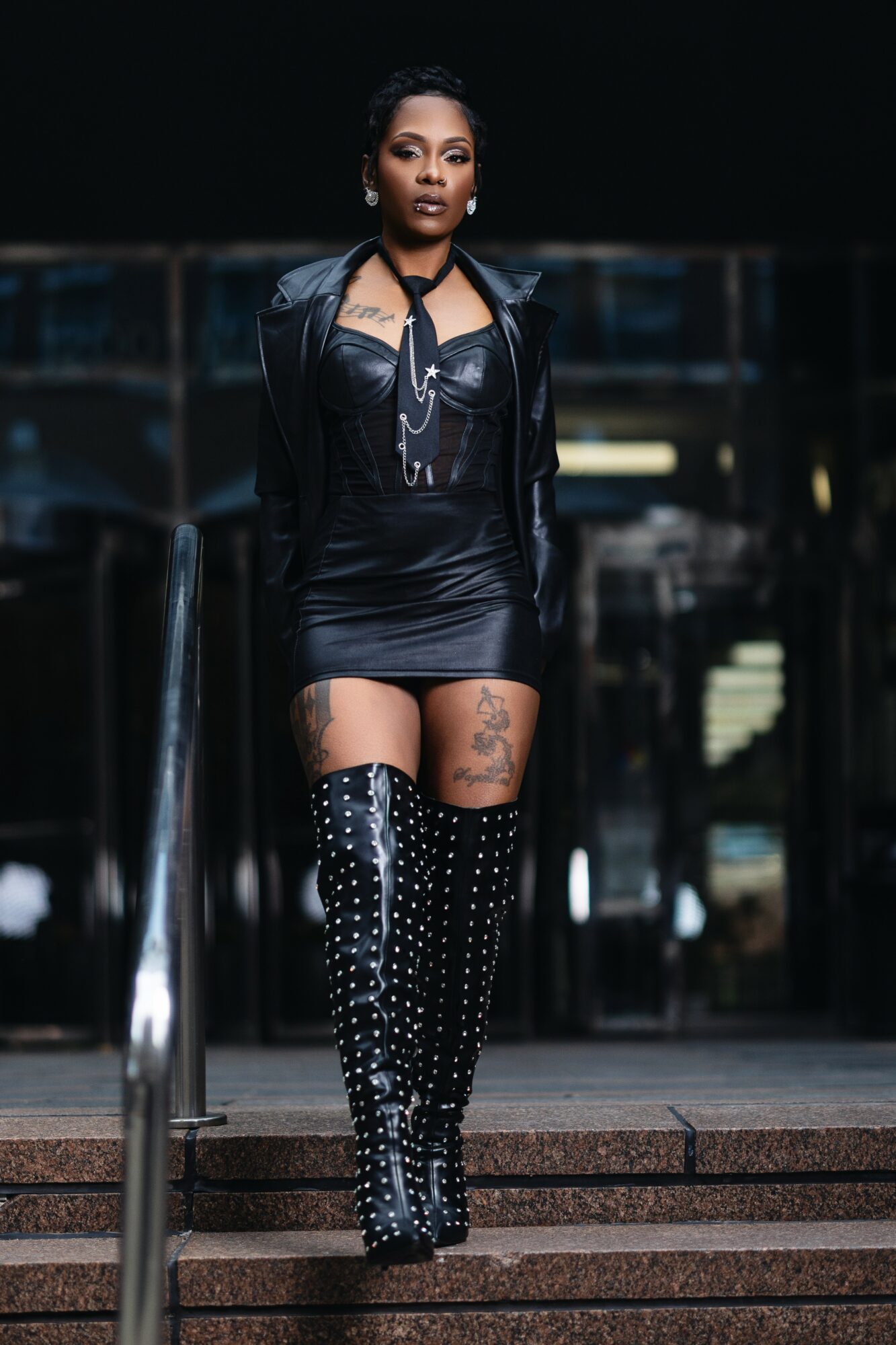 Woman standing outdoors on steps, wearing a black leather jacket, short dress, and thigh-high polka dot boots, with a dark background.