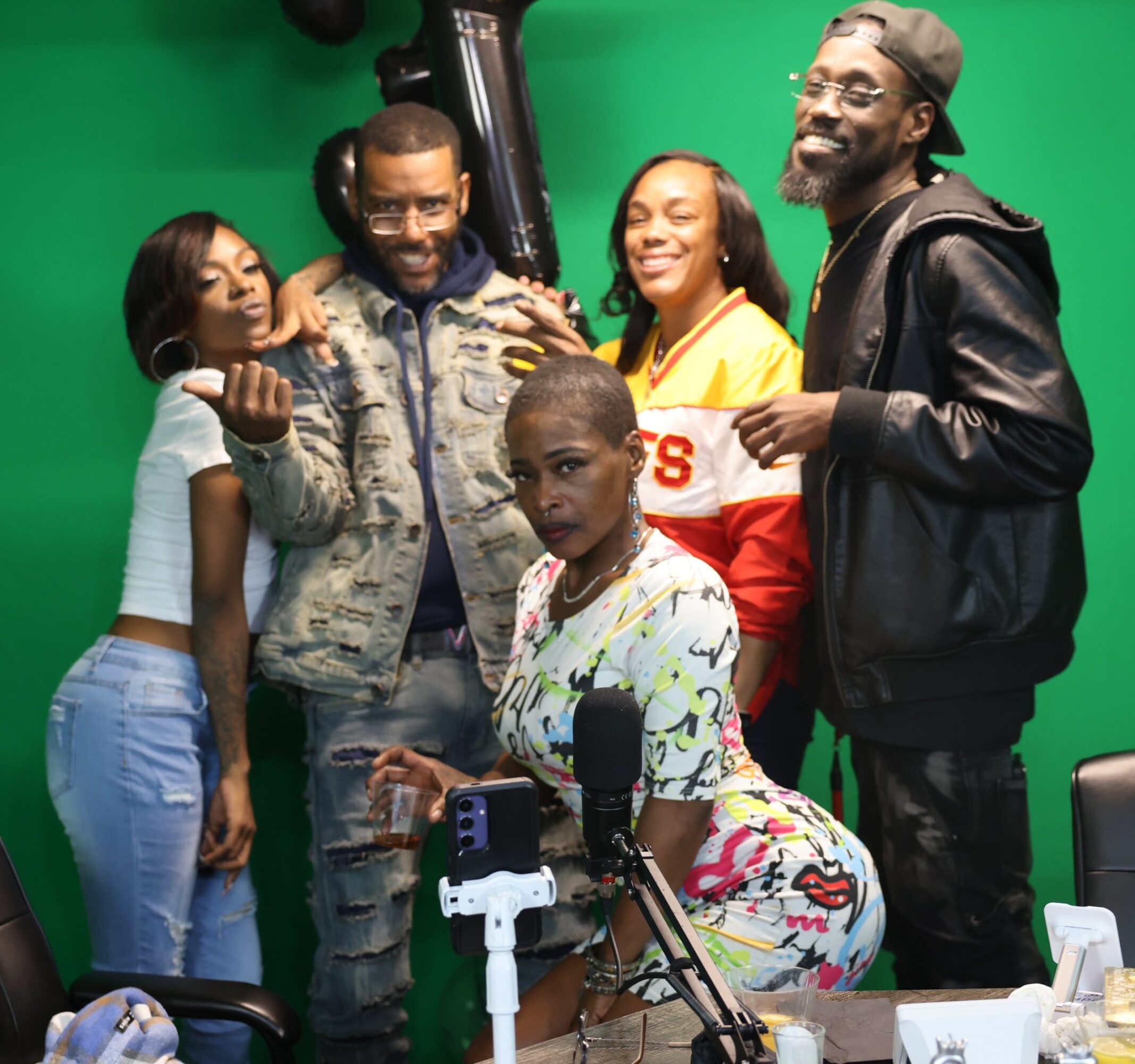 Group of five people smiling and posing in front of a green screen, with recording equipment on a table in front.