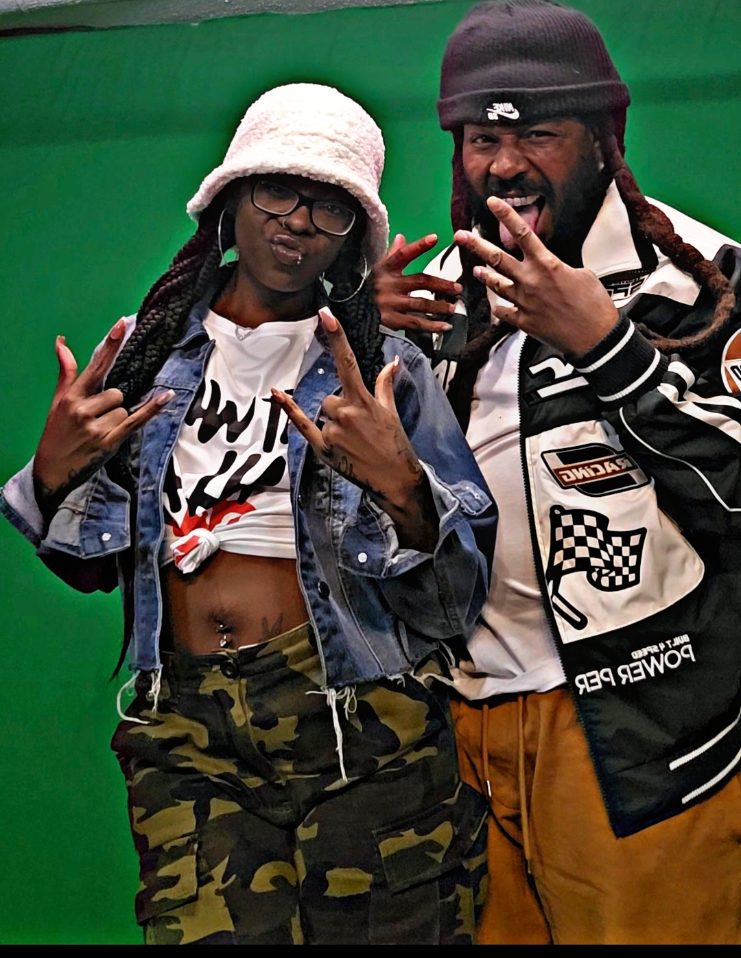 Two people posing with hand gestures, one wearing a bucket hat and camouflage pants, the other in a beanie and jacket, against a green background.