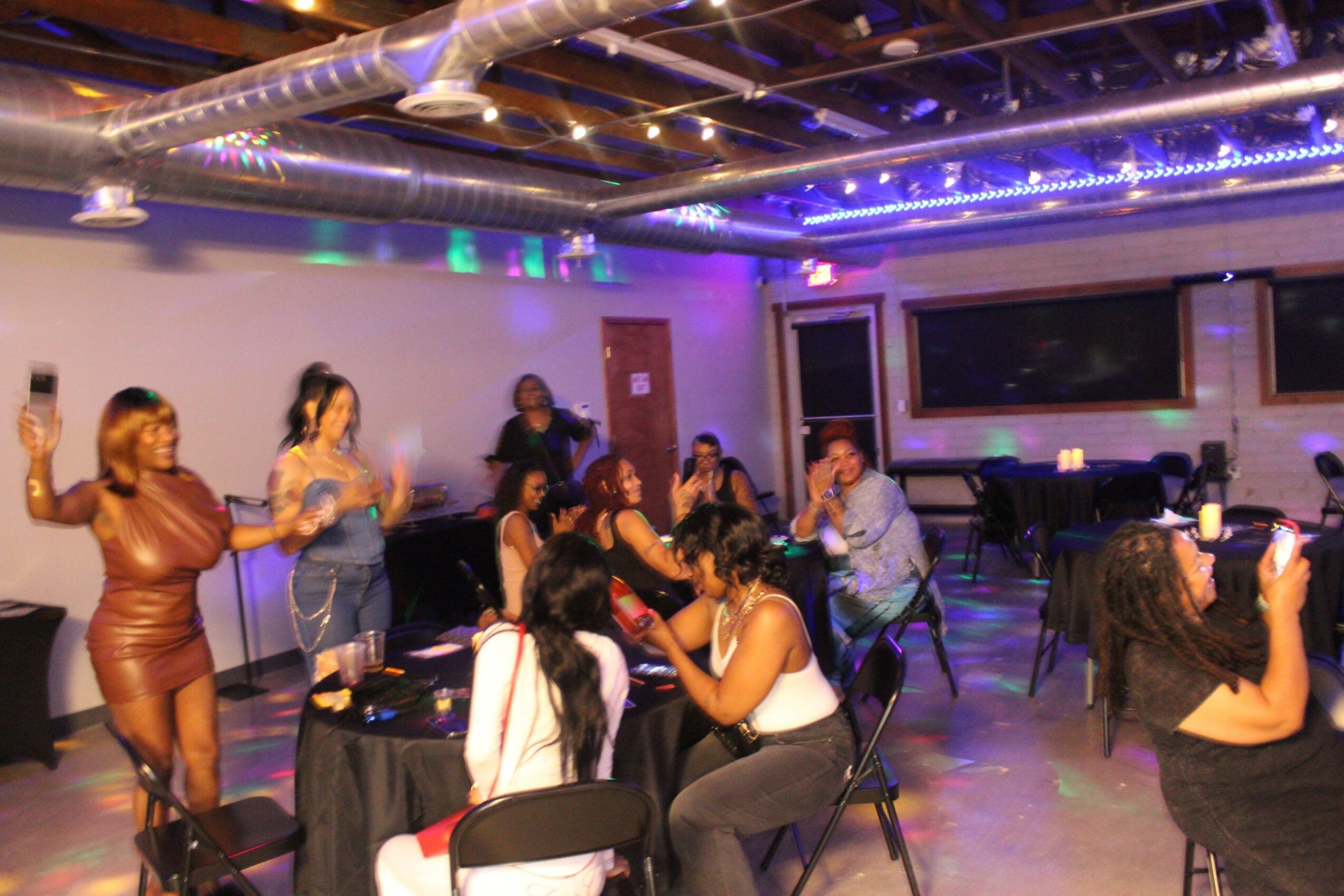 People dancing and socializing in a dimly lit room with tables and a large screen, celebrating or enjoying an event.