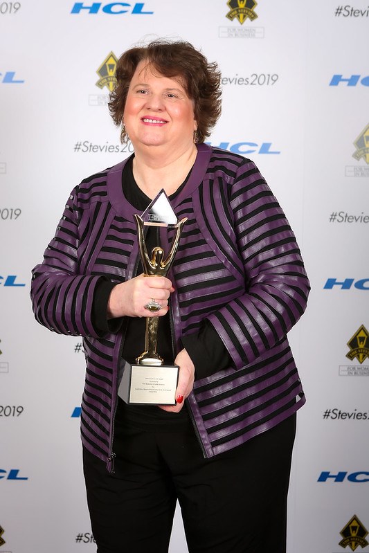 Woman with curly hair smiling, holding an award trophy, standing in front of a backdrop with logos and text.