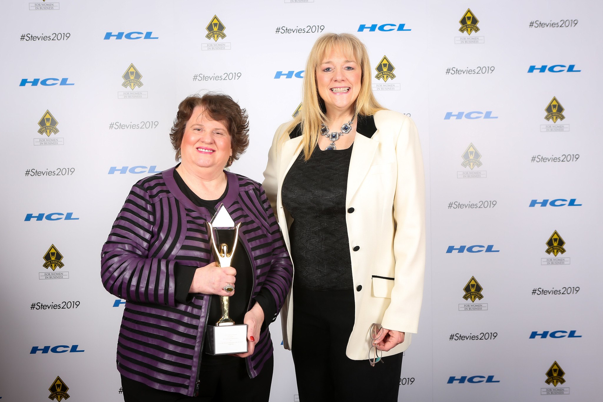 Two women standing in front of a backdrop with logos, one holding a trophy, both smiling.