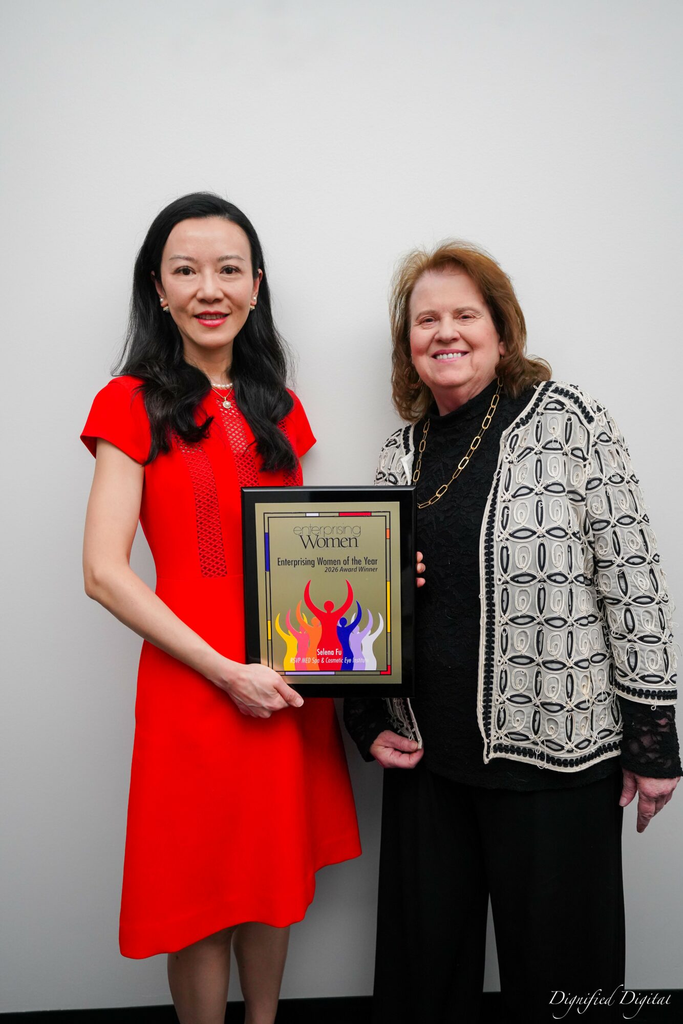 Two women standing together, one holding a framed award, smiling, against a plain white wall.