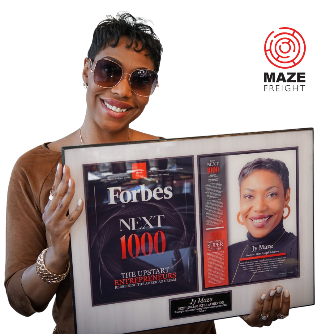 Smiling woman holding a framed magazine cover with Forbes and a portrait, wearing sunglasses and a brown jacket.