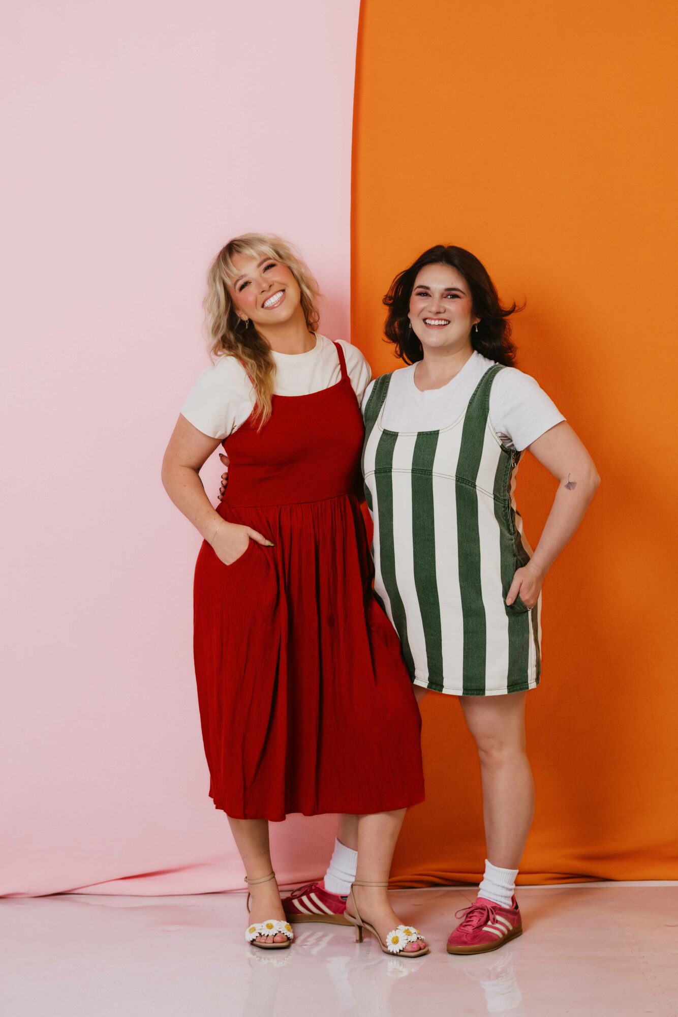 Two women stand side by side, smiling, with a pink and orange background behind them.