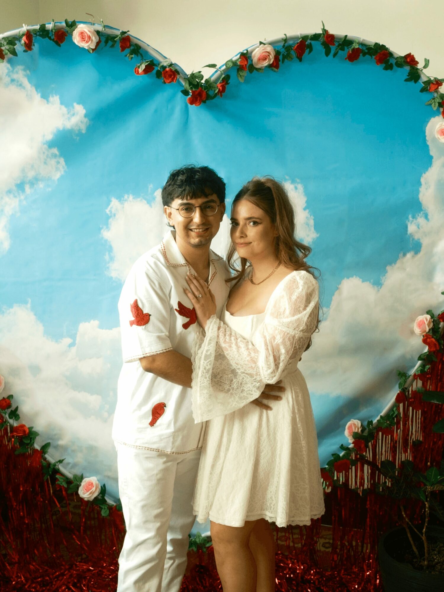 A young man and woman stand together in front of a heart-shaped backdrop with clouds and flowers, smiling at the camera.