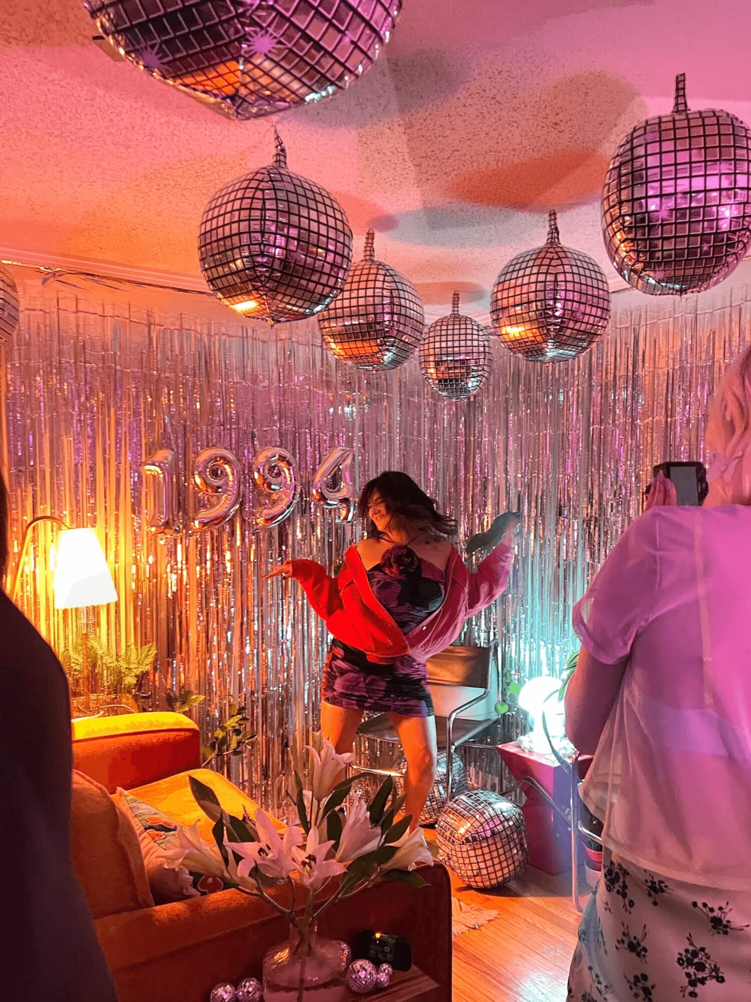 Person dancing in front of shiny hanging disco balls and metallic curtain, with a sofa, lamp, and flowers nearby, celebrating 1999.