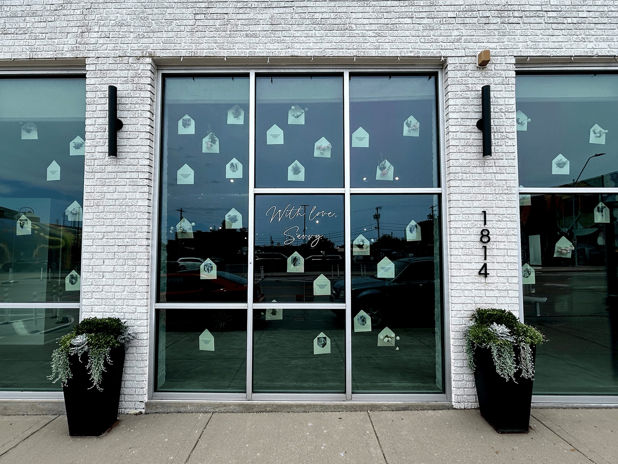Storefront with large glass doors and windows, decorated with small house-shaped stickers, two potted plants outside, and street view reflected in glass.