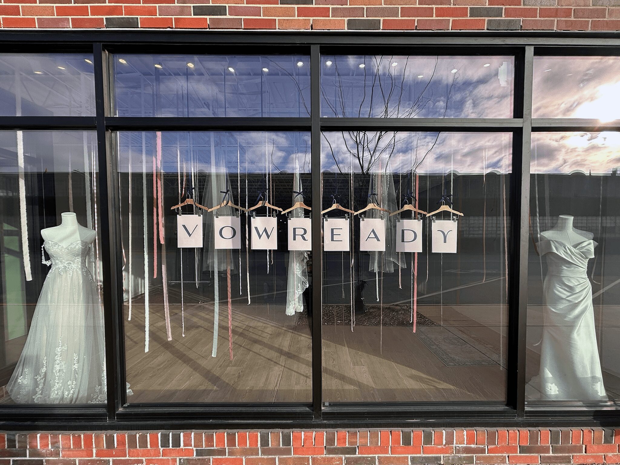 Store window display with wedding dresses and signs spelling 'NOW READY'.