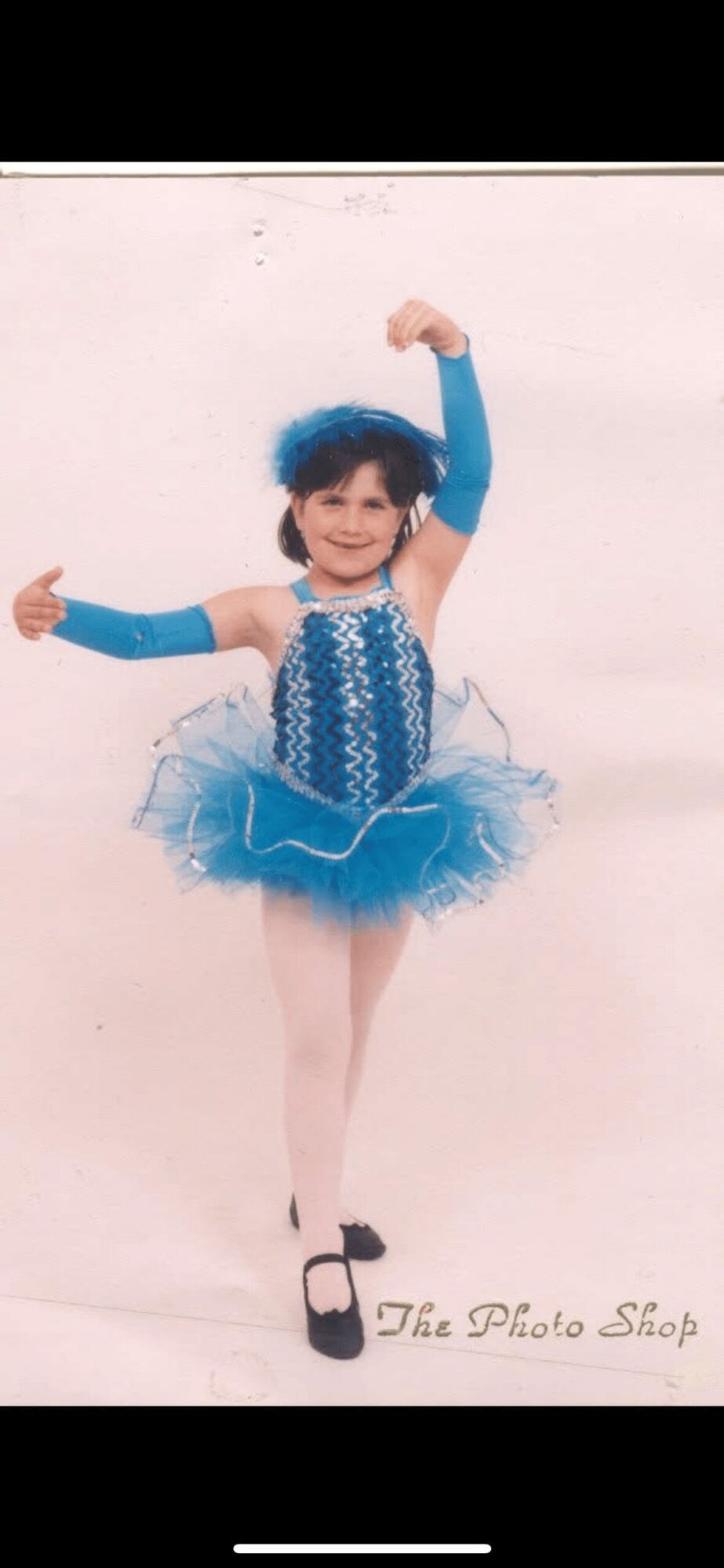 Young girl in a blue ballet costume with tutu, tights, and ballet shoes, smiling with arms raised, standing on a plain background.