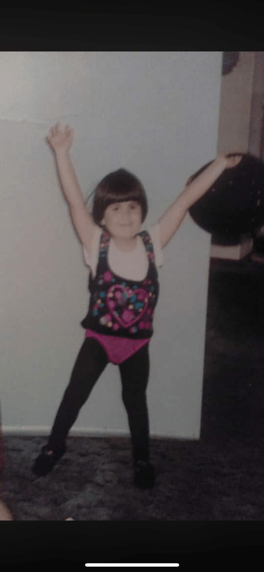 Child with arms raised, wearing a colorful top and black pants, standing indoors against a plain wall.