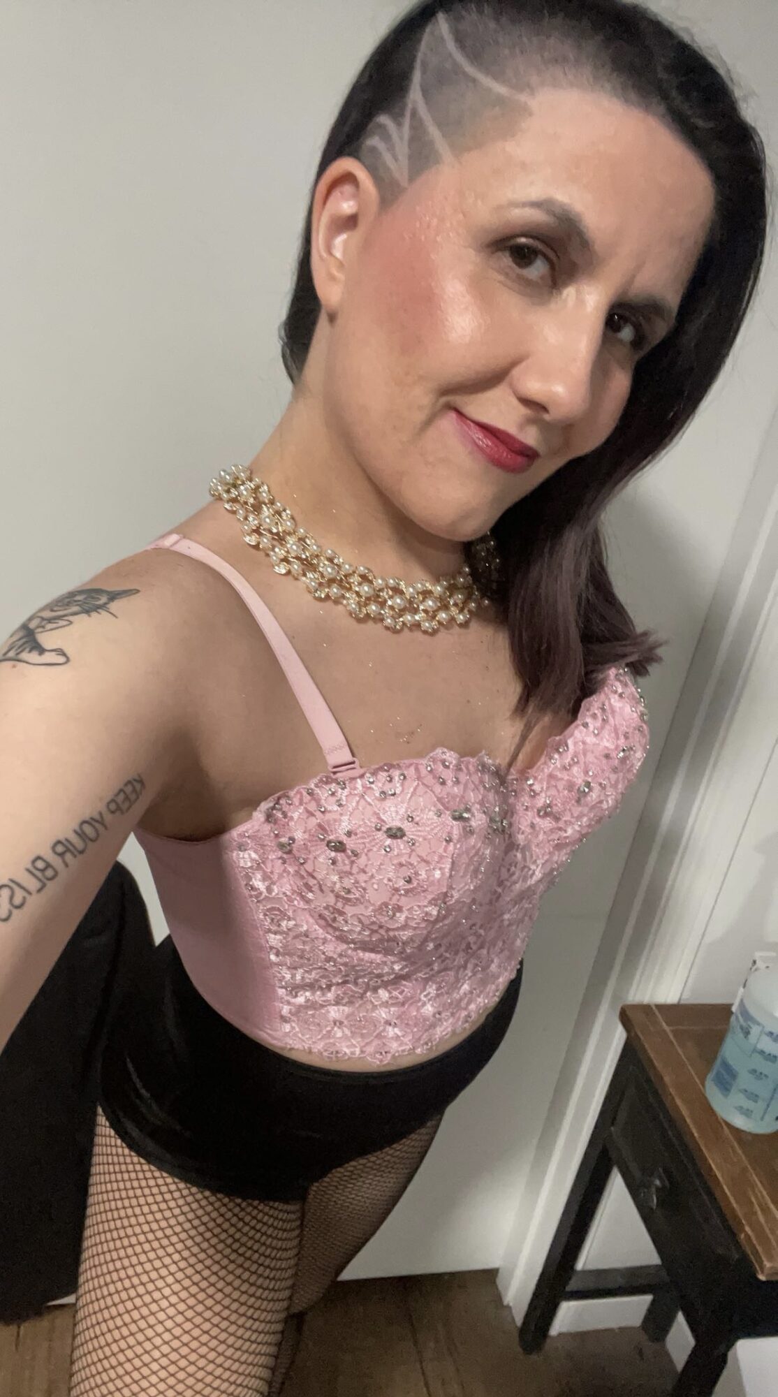 Woman with short dark hair, wearing pink lace top, necklace, and fishnet stockings, smiling at camera.