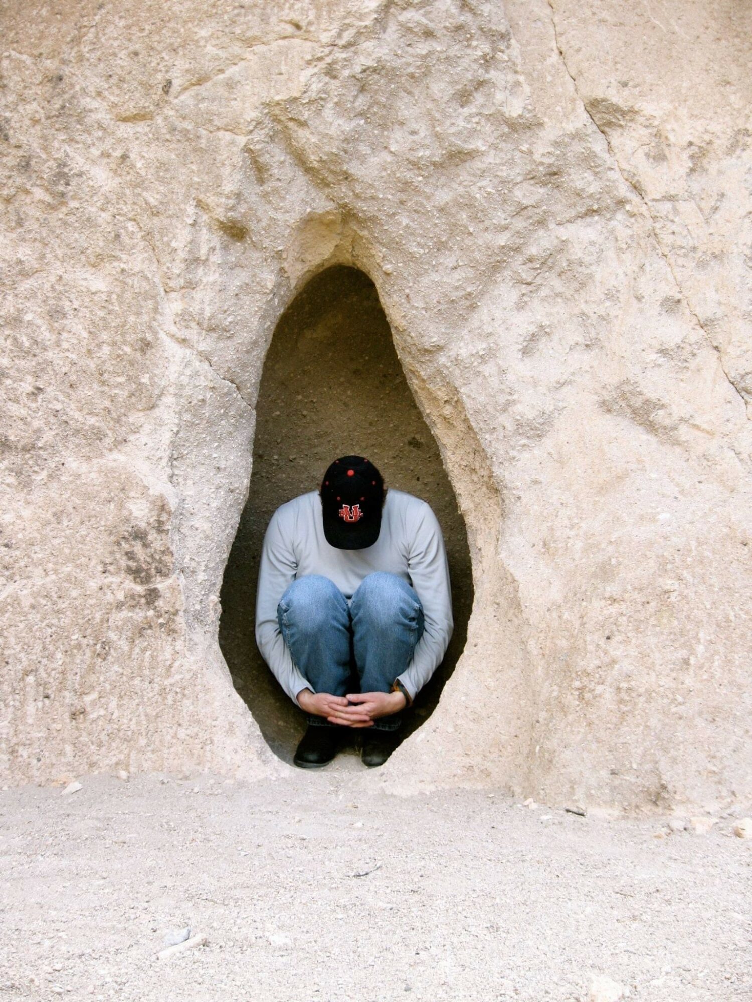 Person crouching inside a small cave opening, wearing a black cap and light blue jeans, with beige rock walls.