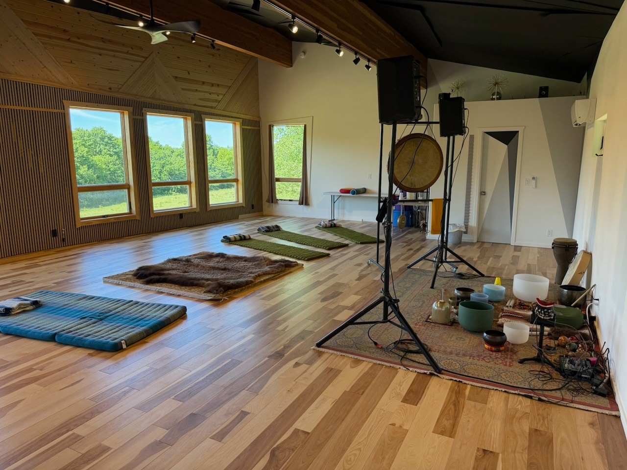 Indoor space with wooden floor, large windows showing greenery, mats on the floor, and a small stage with musical instruments and equipment.
