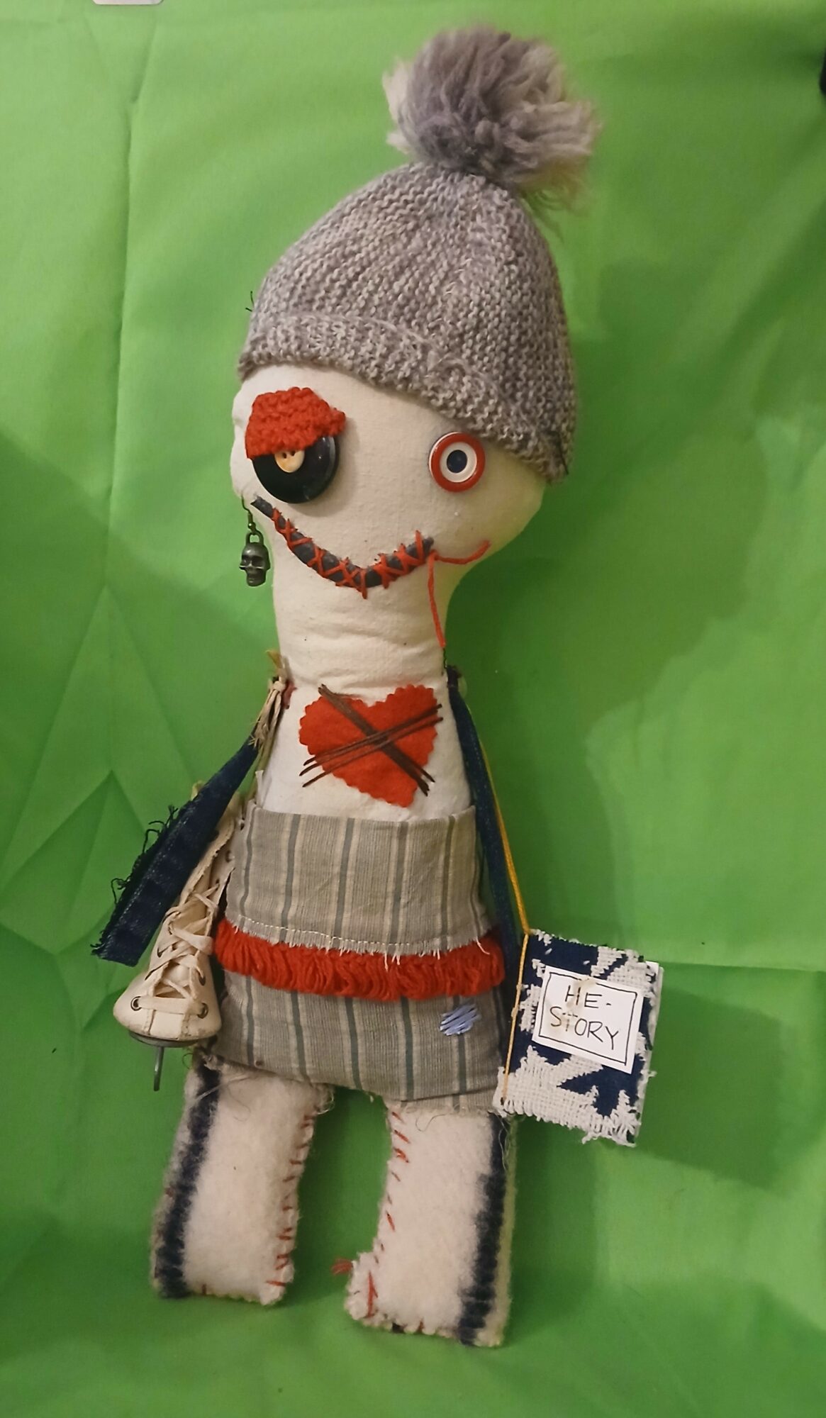 Handmade doll with a gray knit hat, pom-pom, striped clothing, and a patchwork heart, standing against a green background.