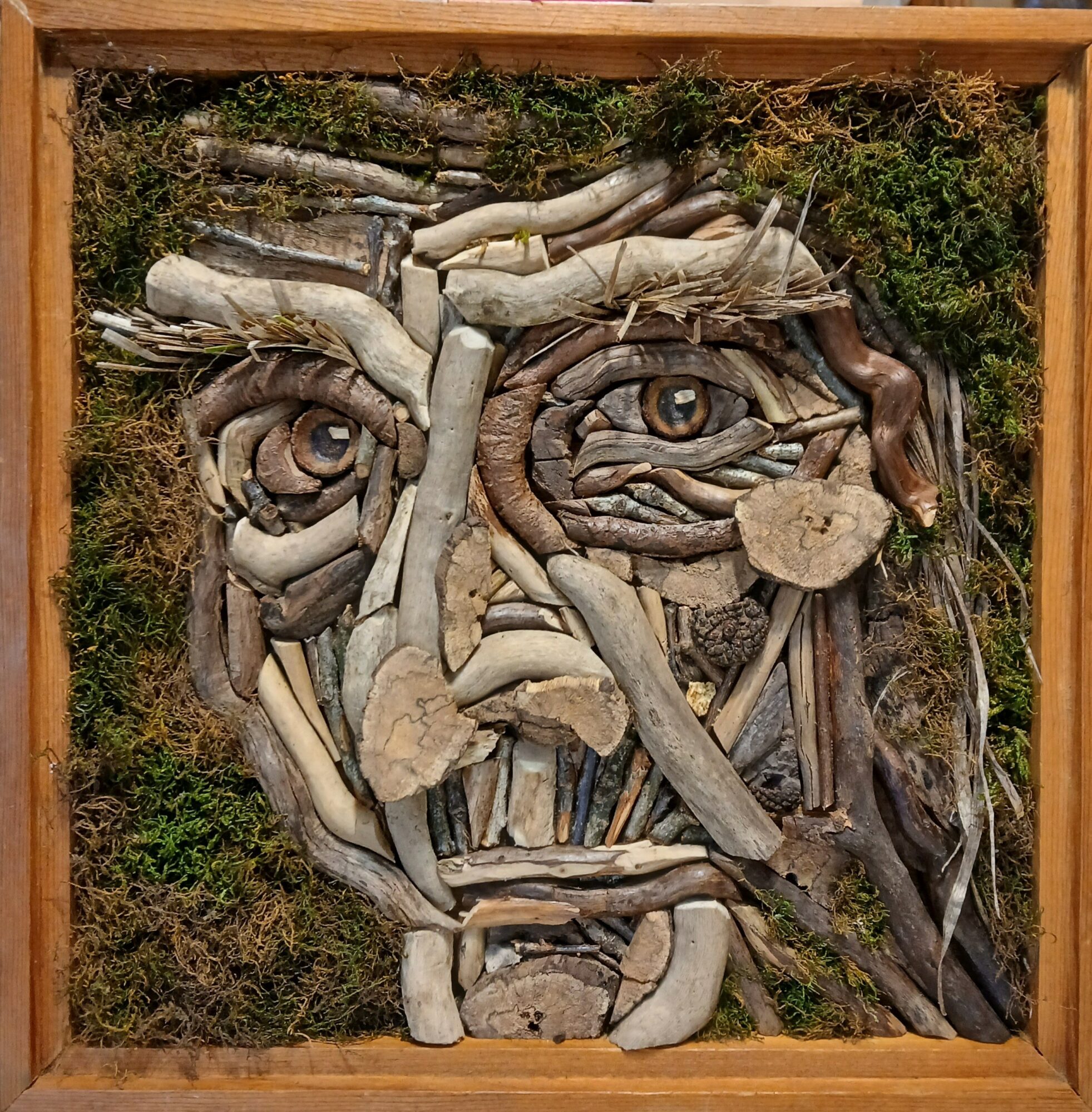 Portrait of a face made from various natural materials like wood and moss, framed in wood, with detailed eyes and facial features.