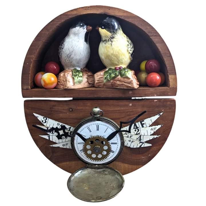 Decorative clock with two bird figurines, fruit, and a clock face with gears, on a wooden background.