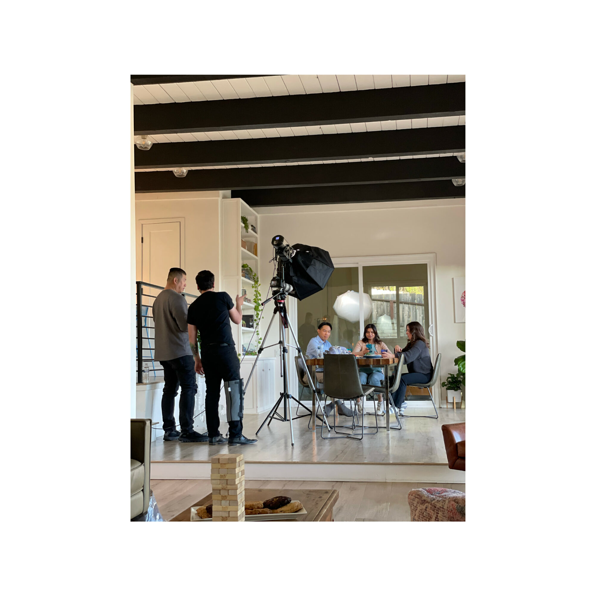 People filming a video or photo shoot in a bright living room with a camera and lighting equipment, four seated at a table.