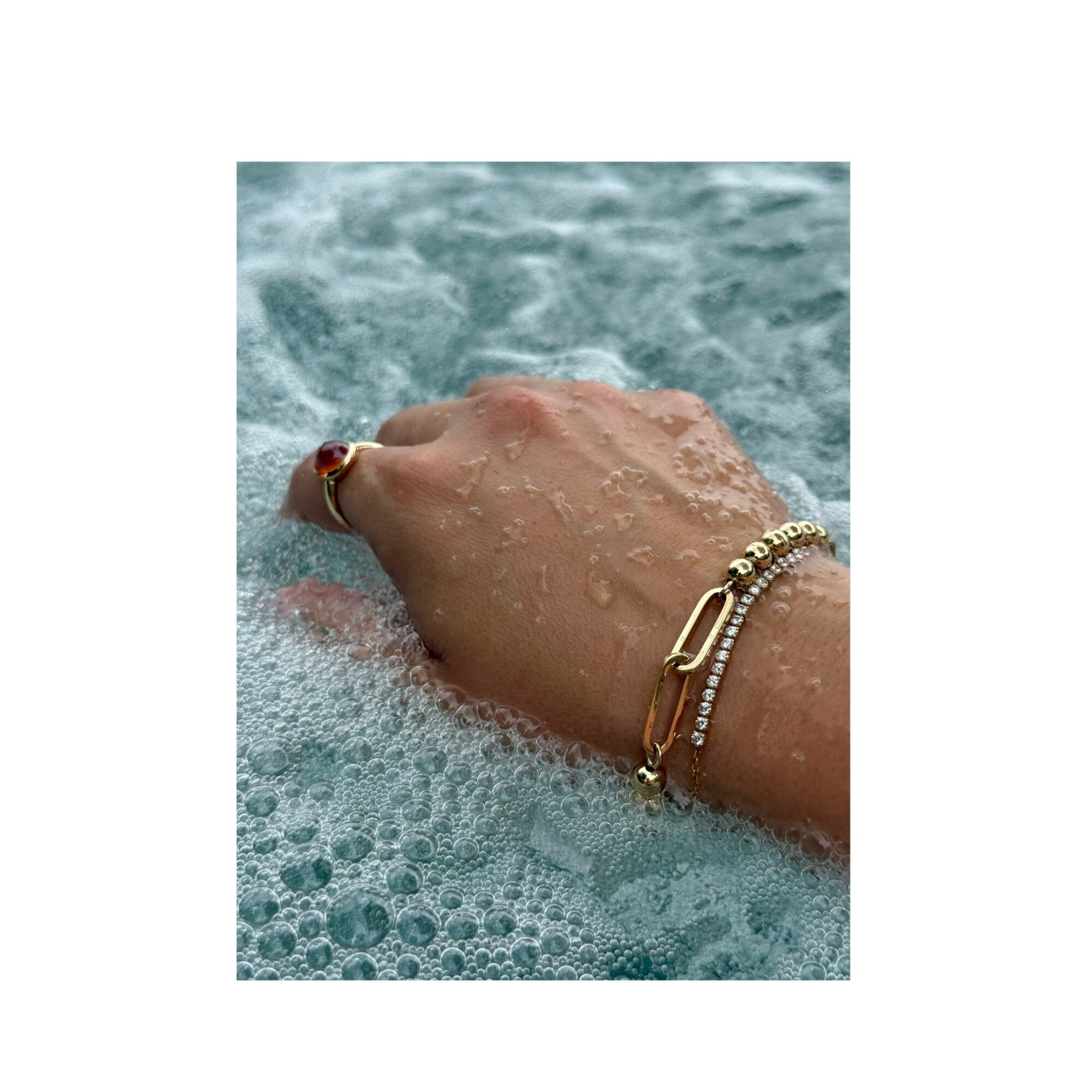 Hand with jewelry submerged in foamy water, possibly at a pool or bath, with bubbles around the hand.