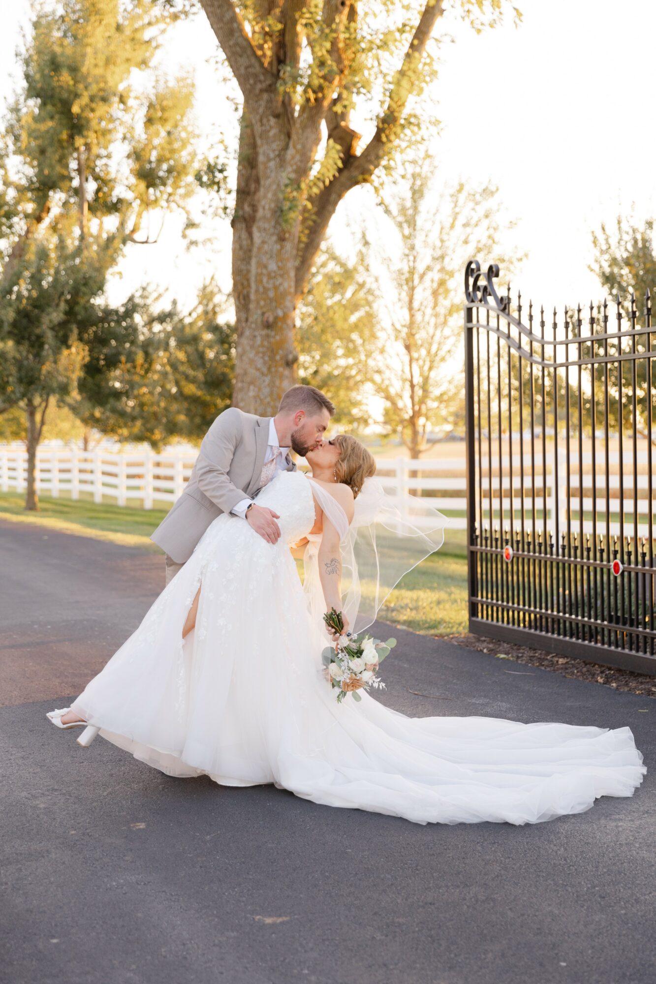 Bride and groom kiss outdoors near a tree and black gate, with sunlight and greenery in background.
