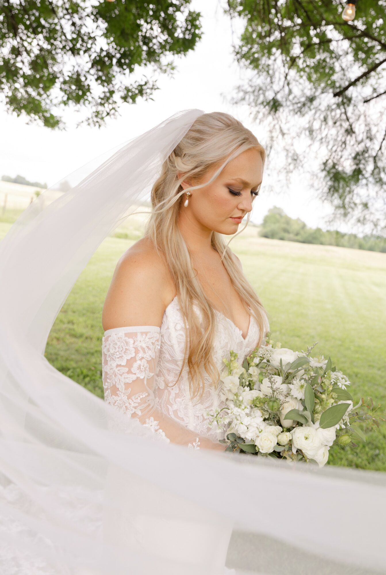 Bride with long blonde hair in a white lace wedding dress holding a bouquet outdoors under trees.
