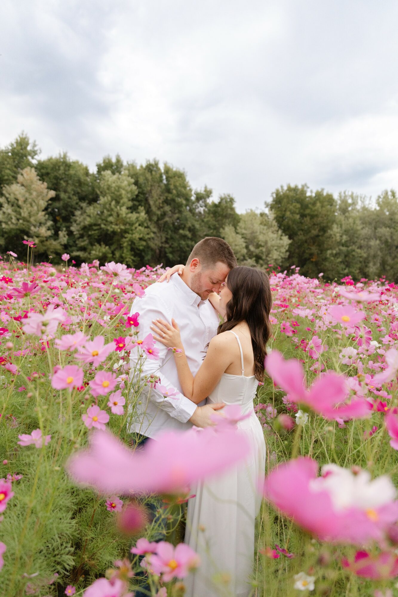 A couple stands close in a pink flower field, embracing and about to kiss, with trees and cloudy sky in background.