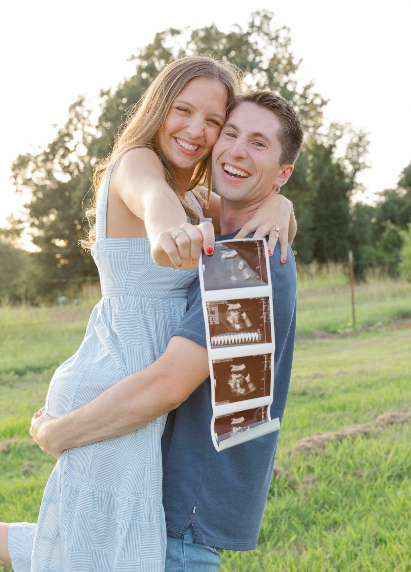 A smiling couple outdoors holding a strip of ultrasound images, with trees and grass in the background.