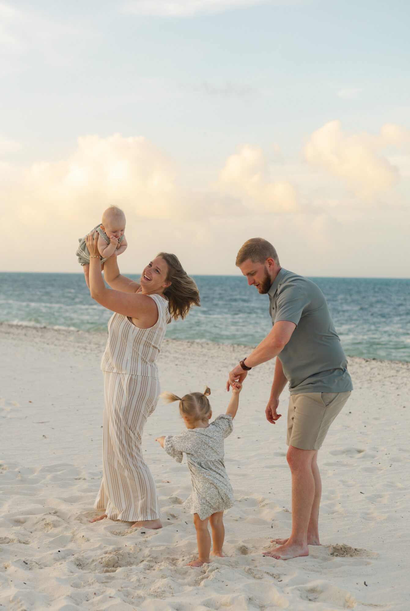 Family of four on a beach, woman holding a baby, child holding father's hand, ocean and sky in background.