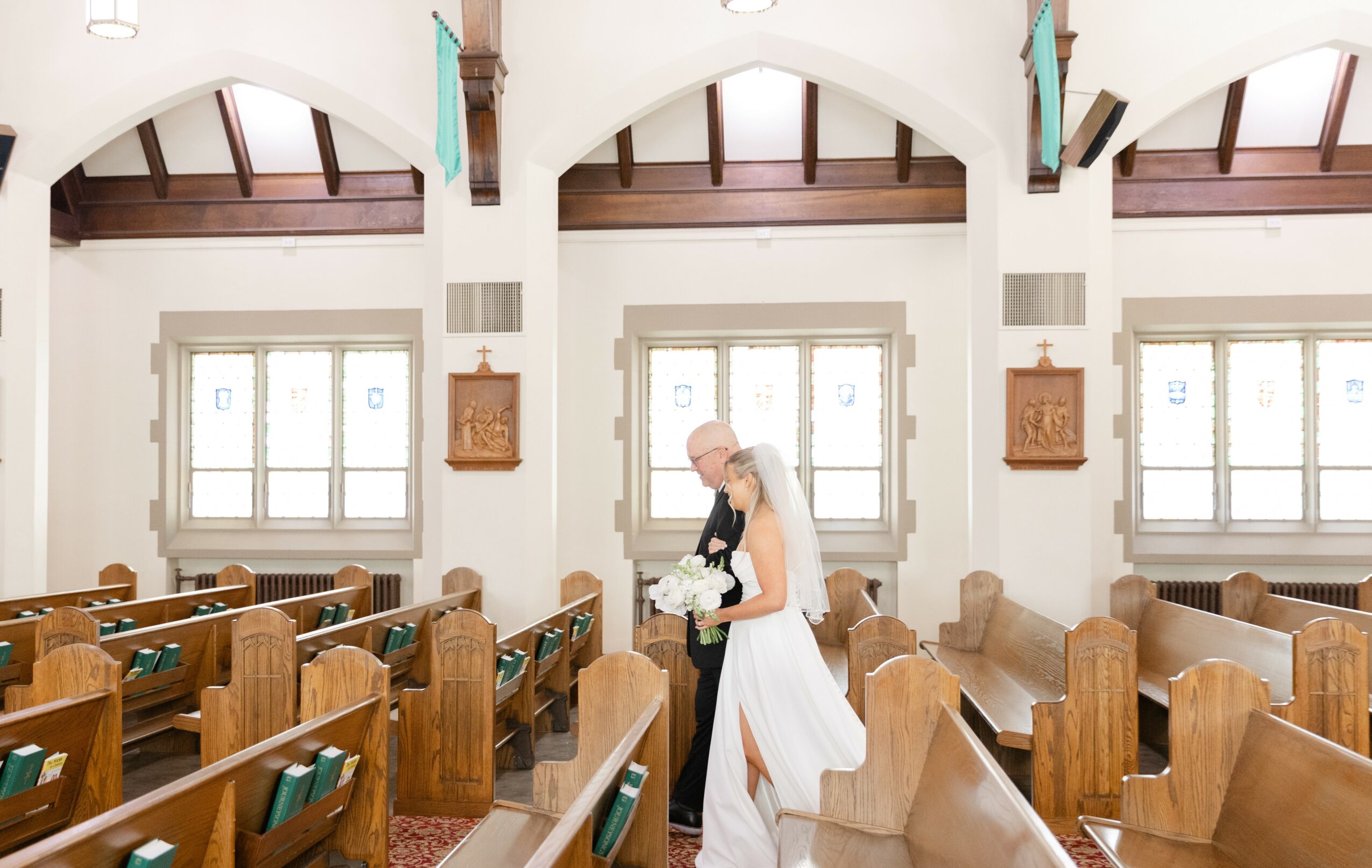 A bride in a white dress holding a bouquet stands inside a church with wooden pews and large windows.