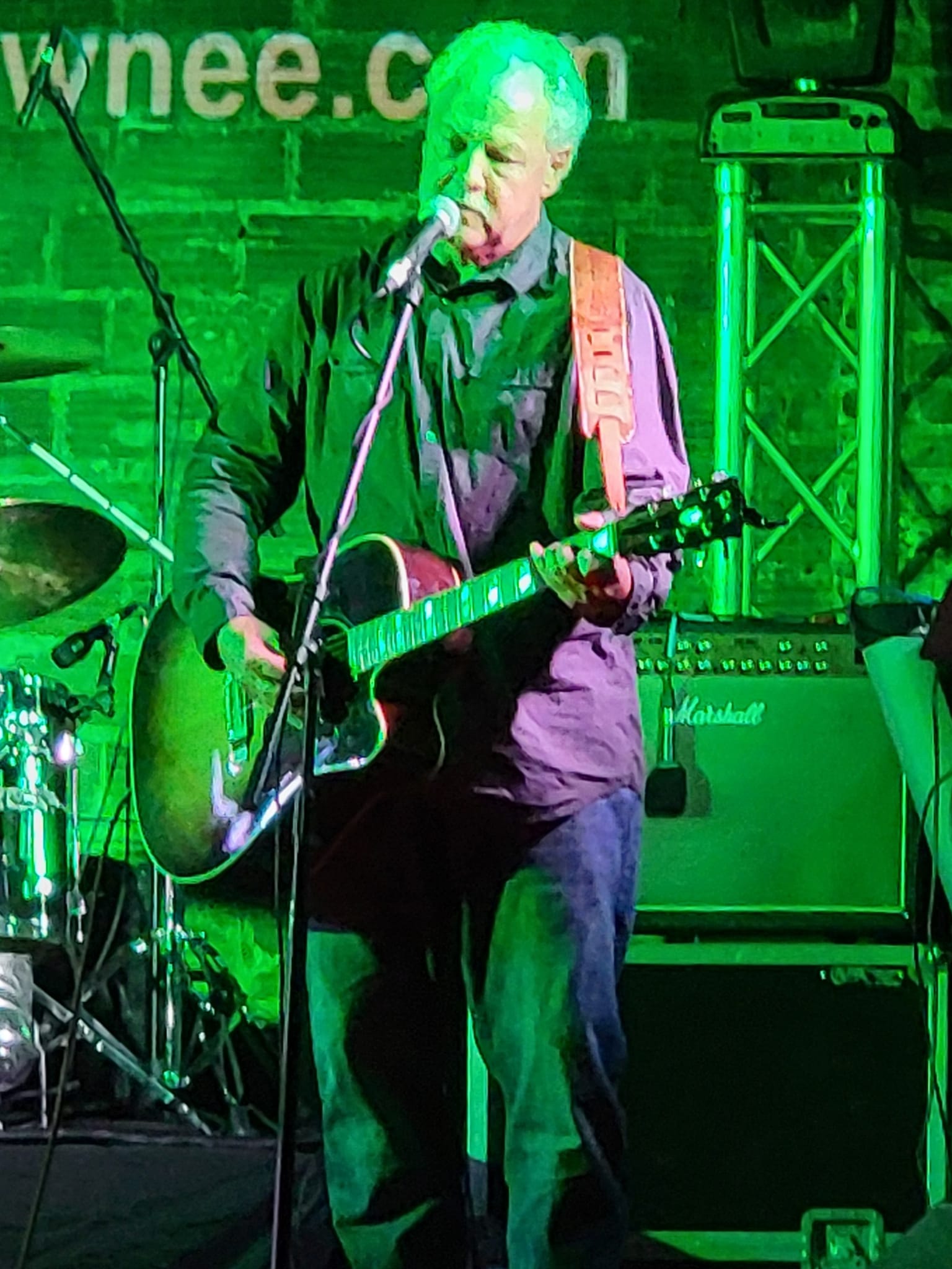 Older man playing acoustic guitar and singing into microphone on stage with musical equipment, green lighting, and brick wall background.