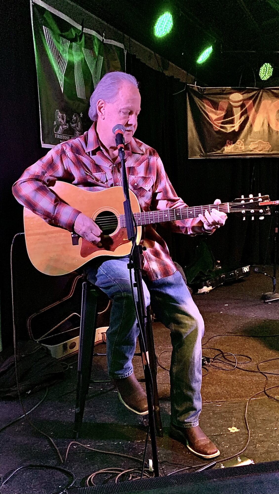 Man playing acoustic guitar and singing on stage with green and orange lighting, seated on a stool, wearing a plaid shirt.