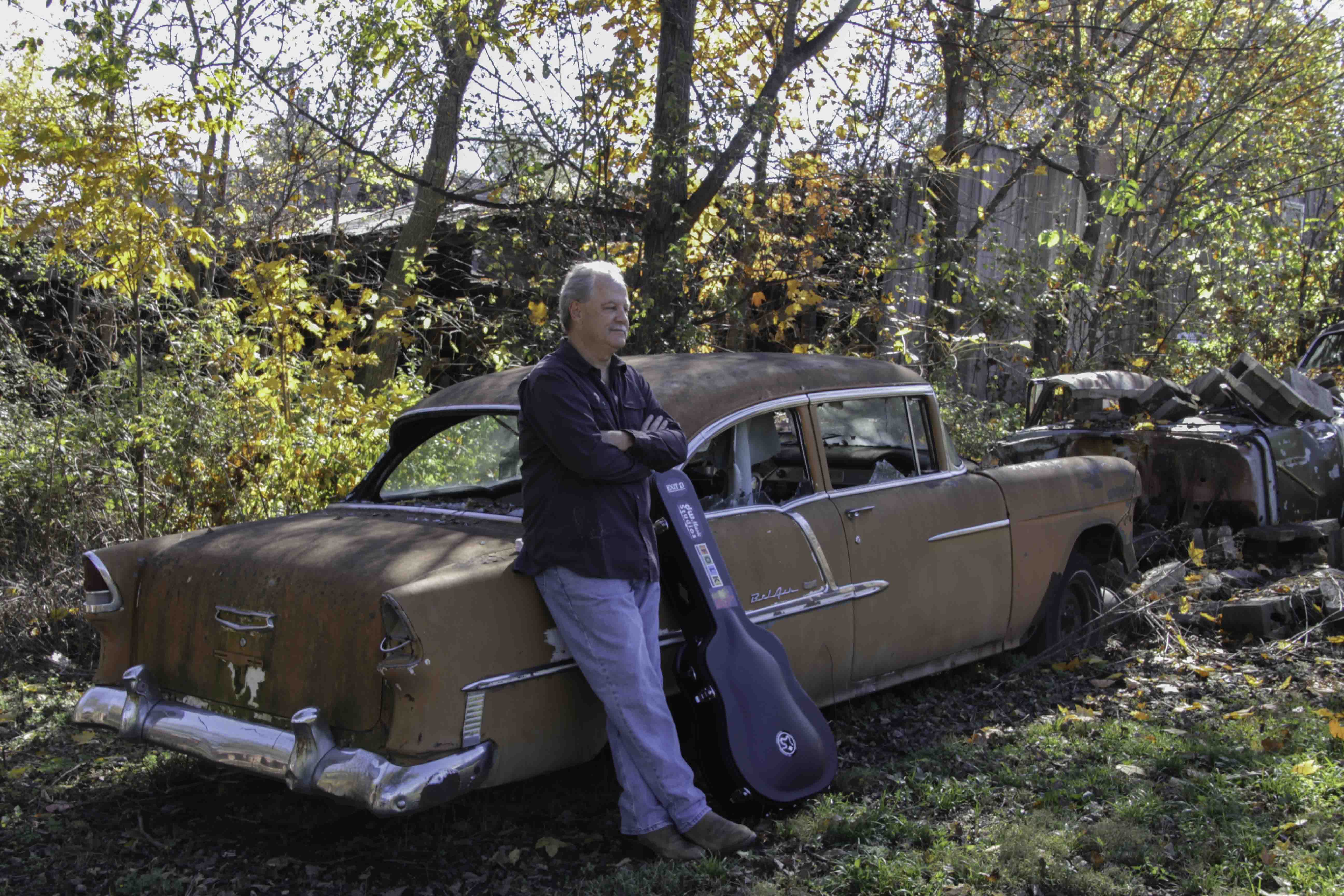 Man standing next to a rusty, vintage car with a guitar case in a wooded area during autumn.
