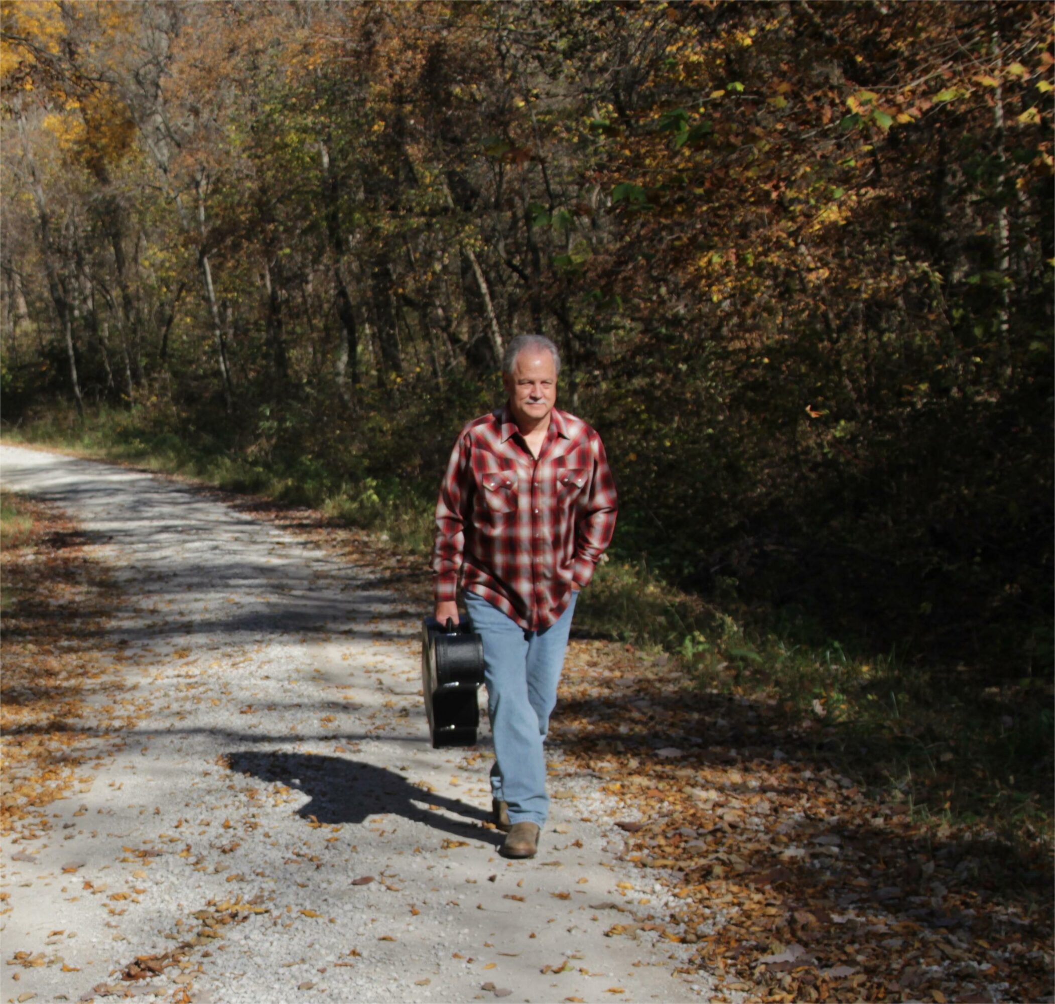 Person walking on a dirt path carrying a black bag, surrounded by trees with autumn leaves.
