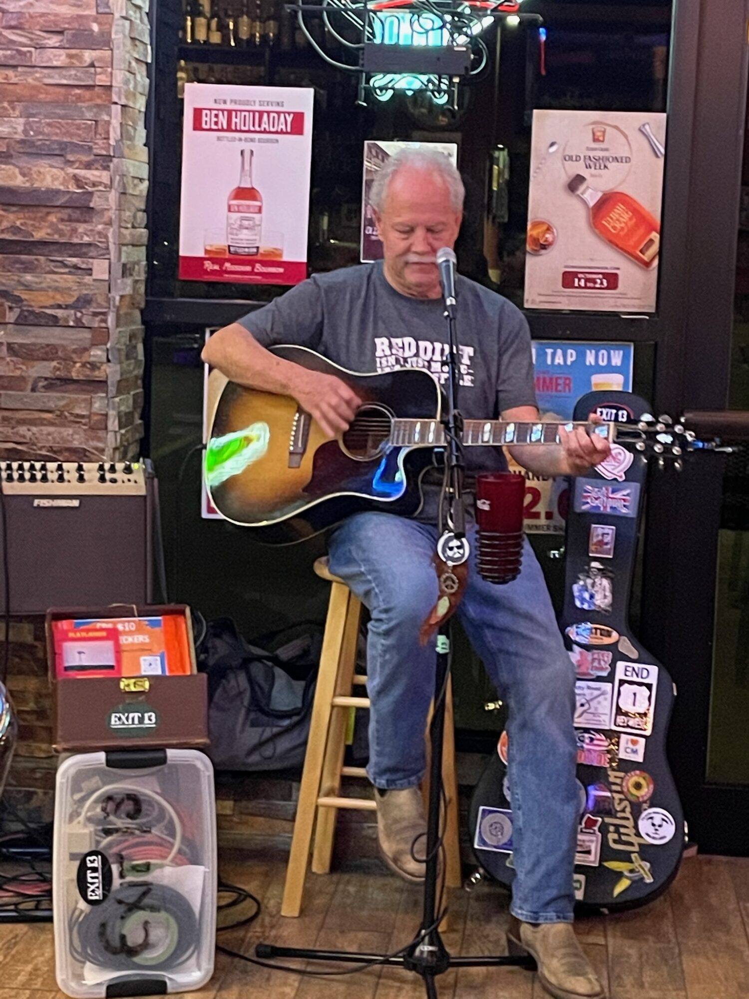Man playing guitar and singing into microphone in a bar, with guitar case and promotional posters nearby.