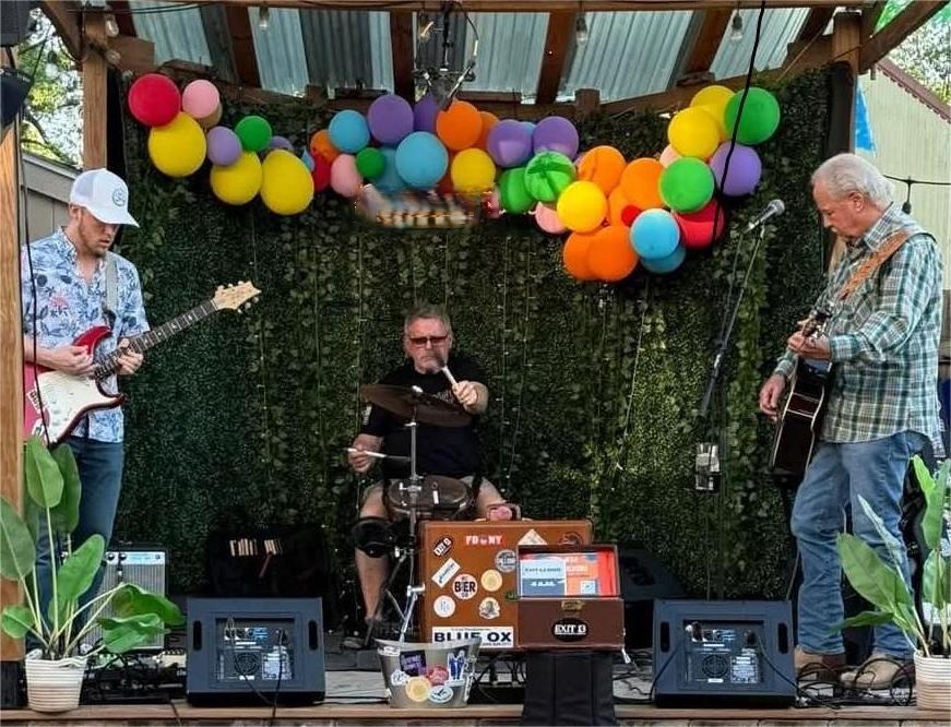 Three musicians perform on a stage decorated with colorful balloons and green foliage backdrop, with two playing guitars and one singing.