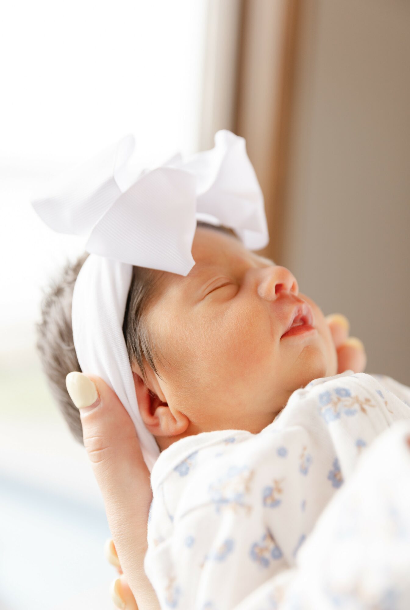Newborn baby with a white bow headband, sleeping peacefully, held by a person, in a bright room.