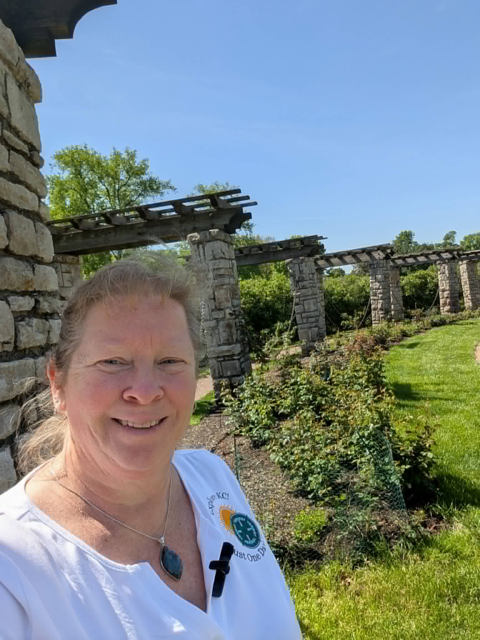 Woman with light hair smiling outdoors near stone ruins and greenery, wearing a white top and a necklace.