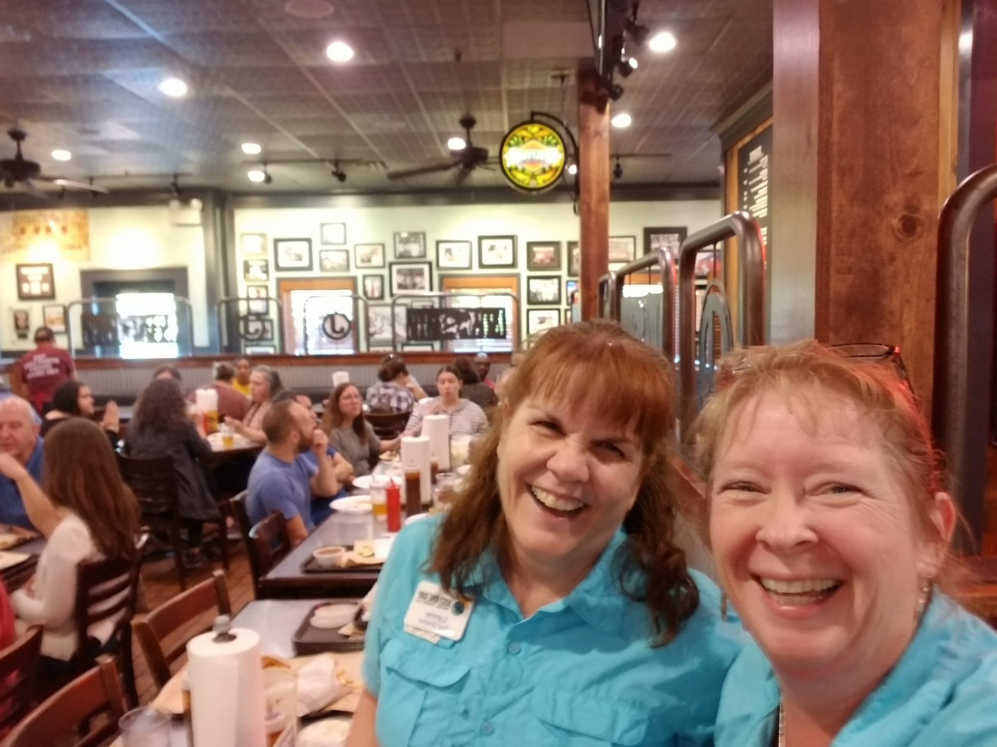 Two women smiling in a busy restaurant with many diners and framed pictures on the wall behind them.