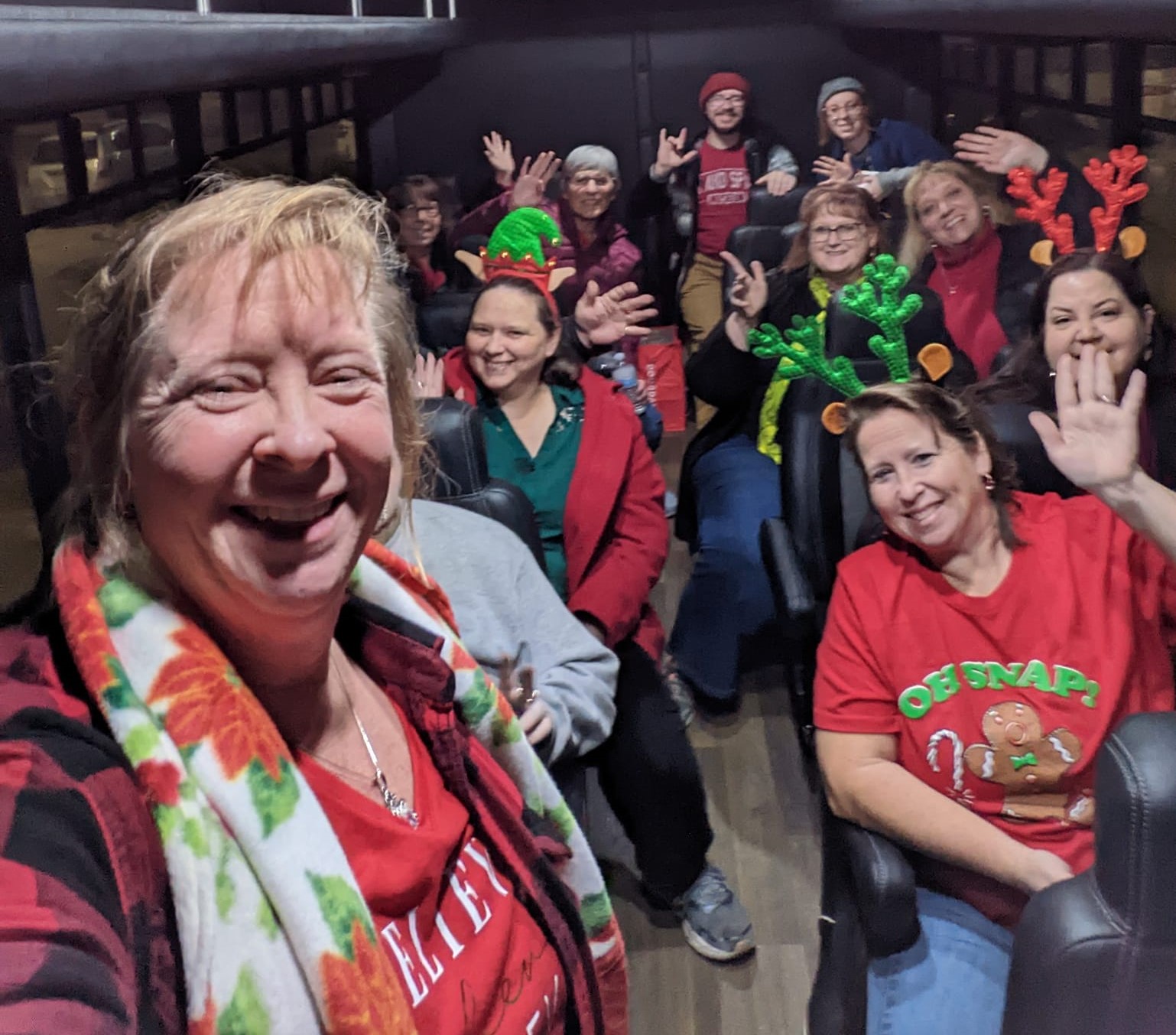 Group of people on a bus, some wearing Christmas accessories, smiling and waving, with a woman in the foreground taking a selfie.