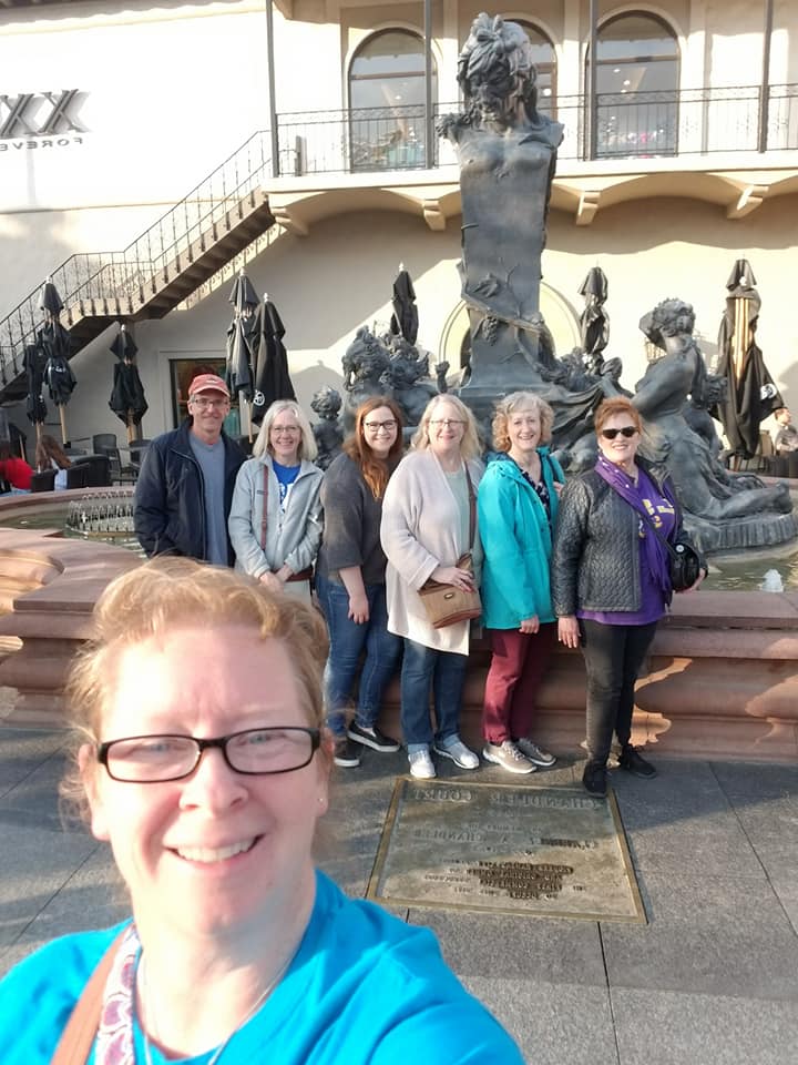 Group of seven people standing in front of a fountain with sculptures, smiling outdoors, with a building and stairs in the background.