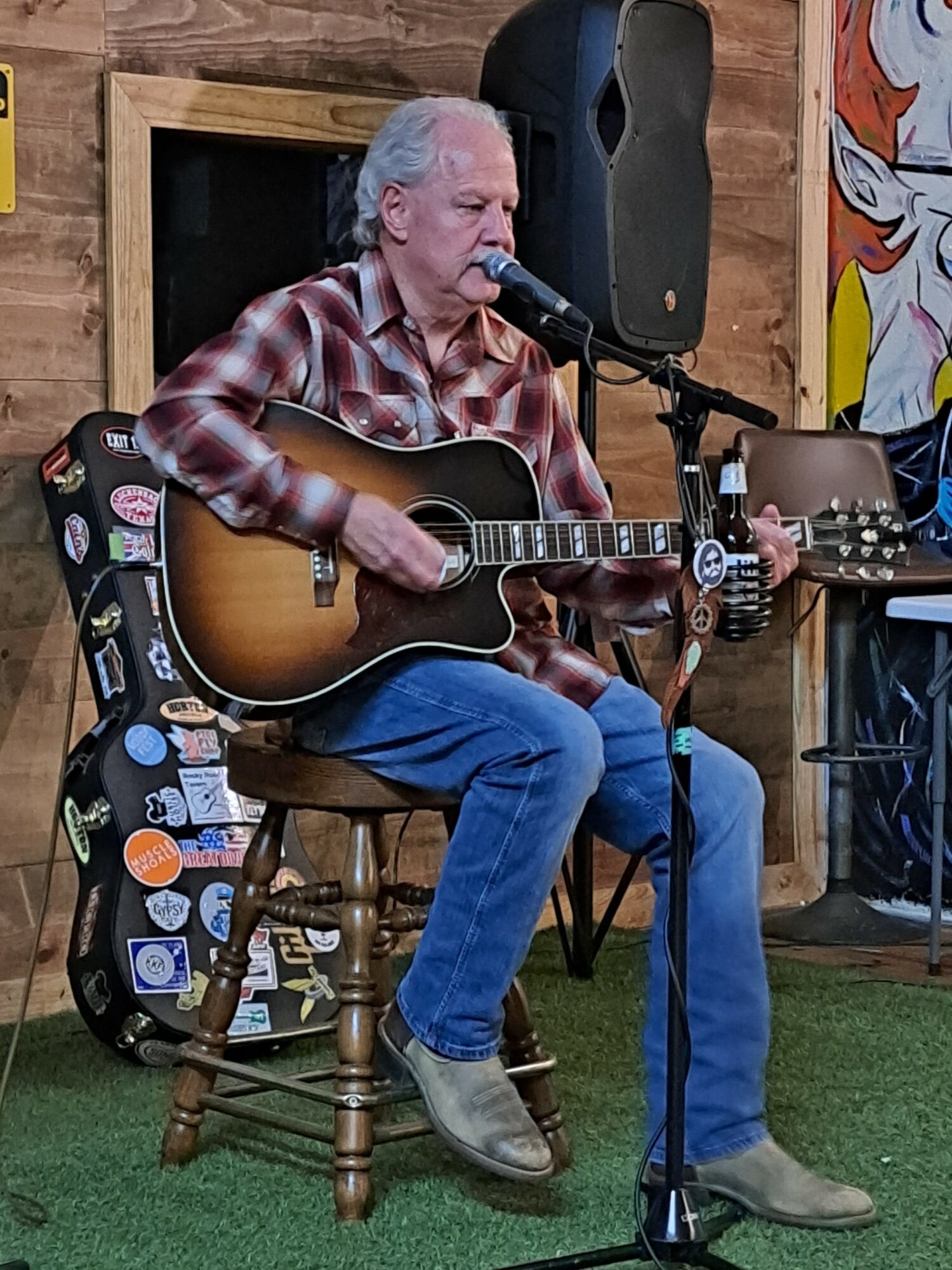 Older man with gray hair playing an acoustic guitar and singing into a microphone in a cozy room.