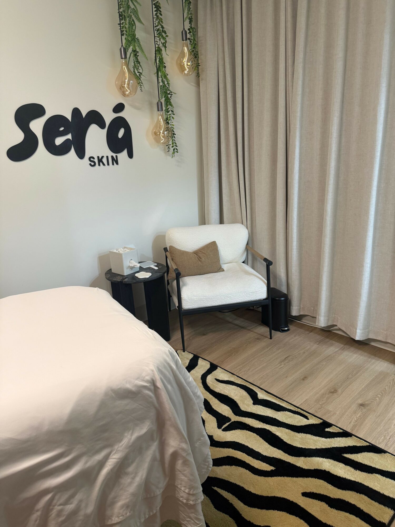 Upright view of a room with a white chair, black side table, zebra-patterned rug, and beige curtains.