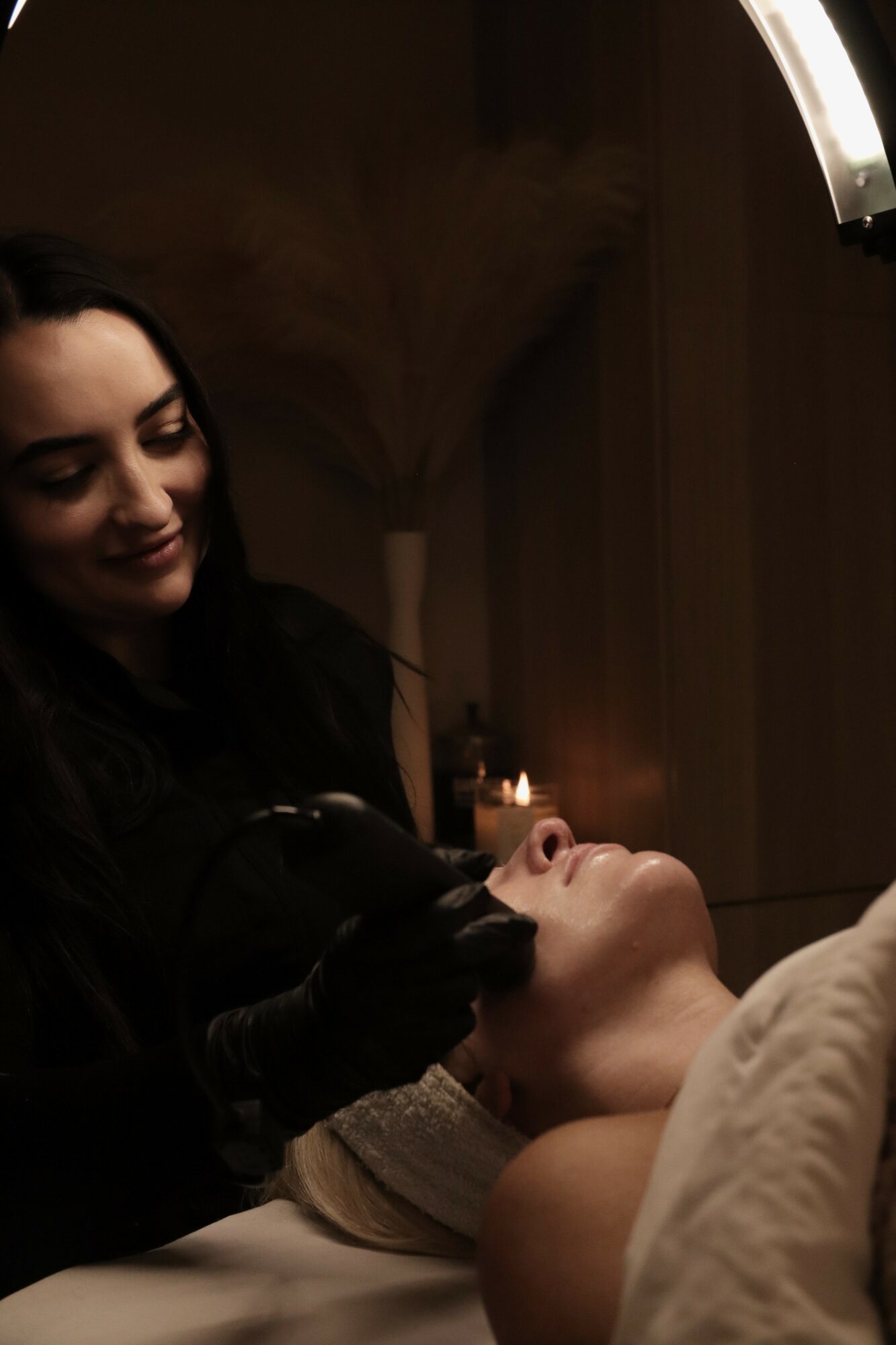 A woman receives a facial treatment in a dimly lit room with candles, smiling gently.