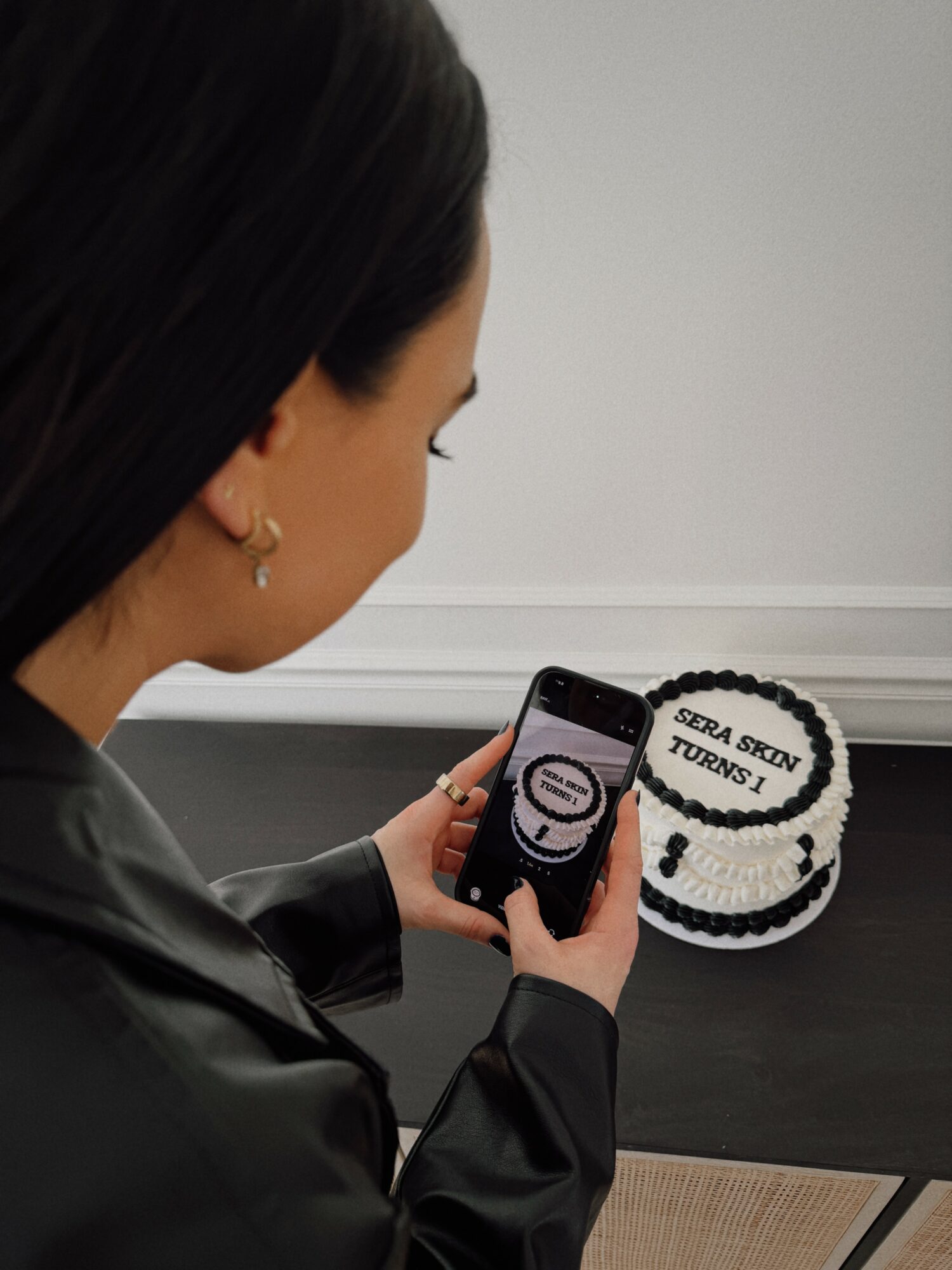 Woman with dark hair and earrings looks at her phone near a cake with writing, against a plain wall.