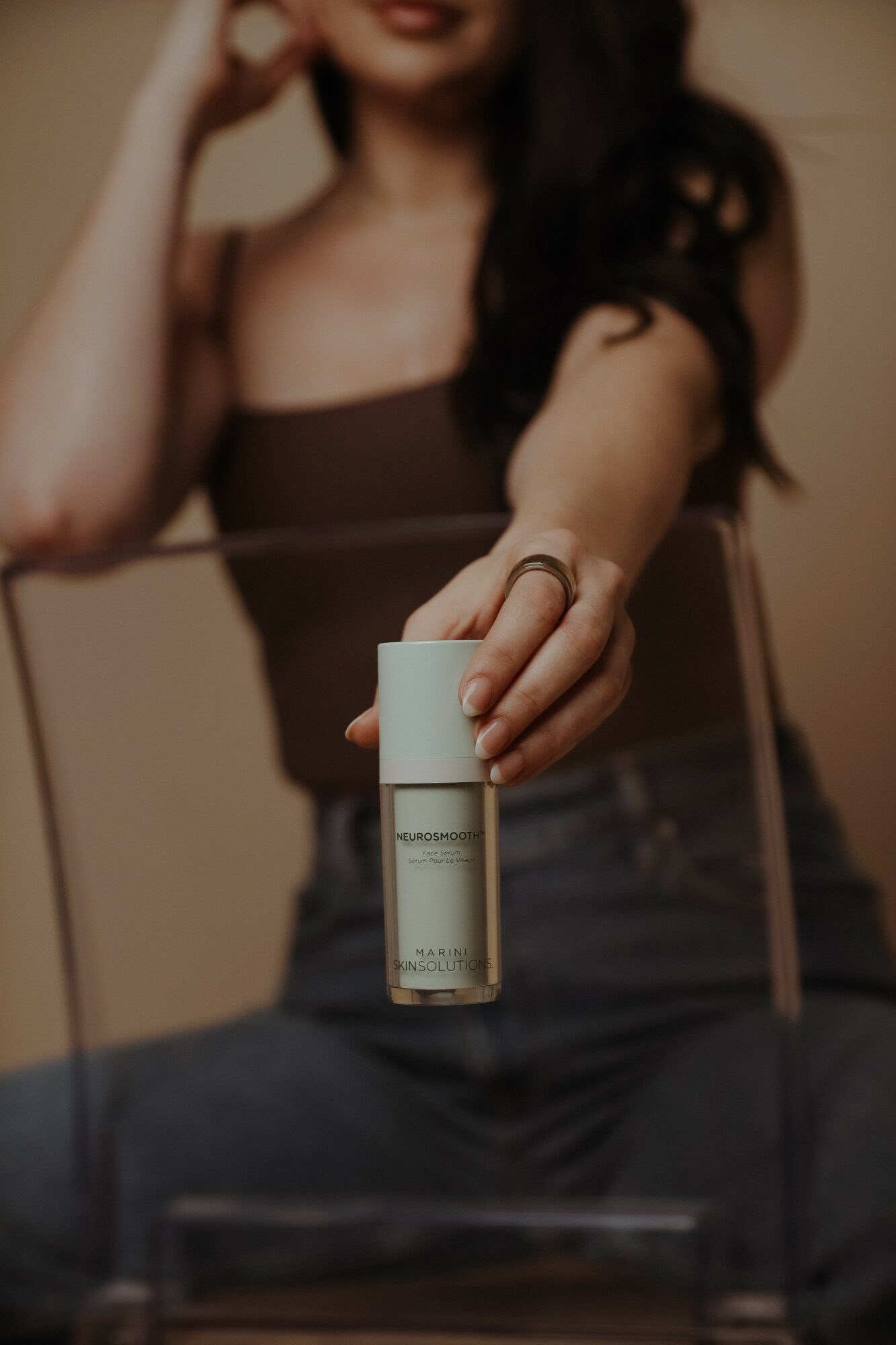 Woman holding a small white bottle towards the camera, sitting on a chair, with blurred background.